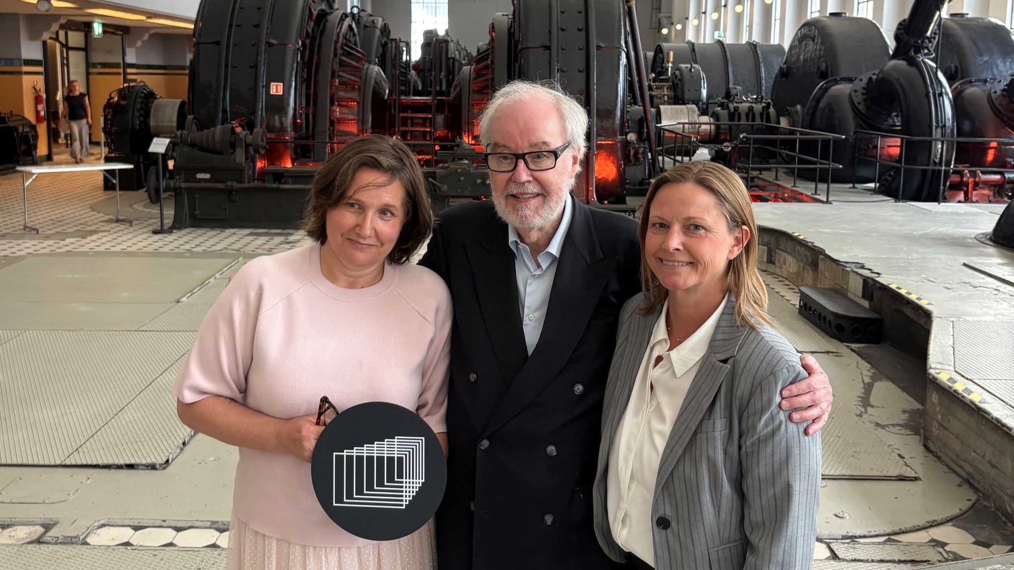 tre personer står i verkstedhall med plakett med European Film Academy logo