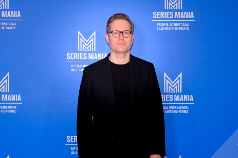 foto. mann ser inn i kamera og står foran en blå fotovegg med flere logoer hvor det står series mania.