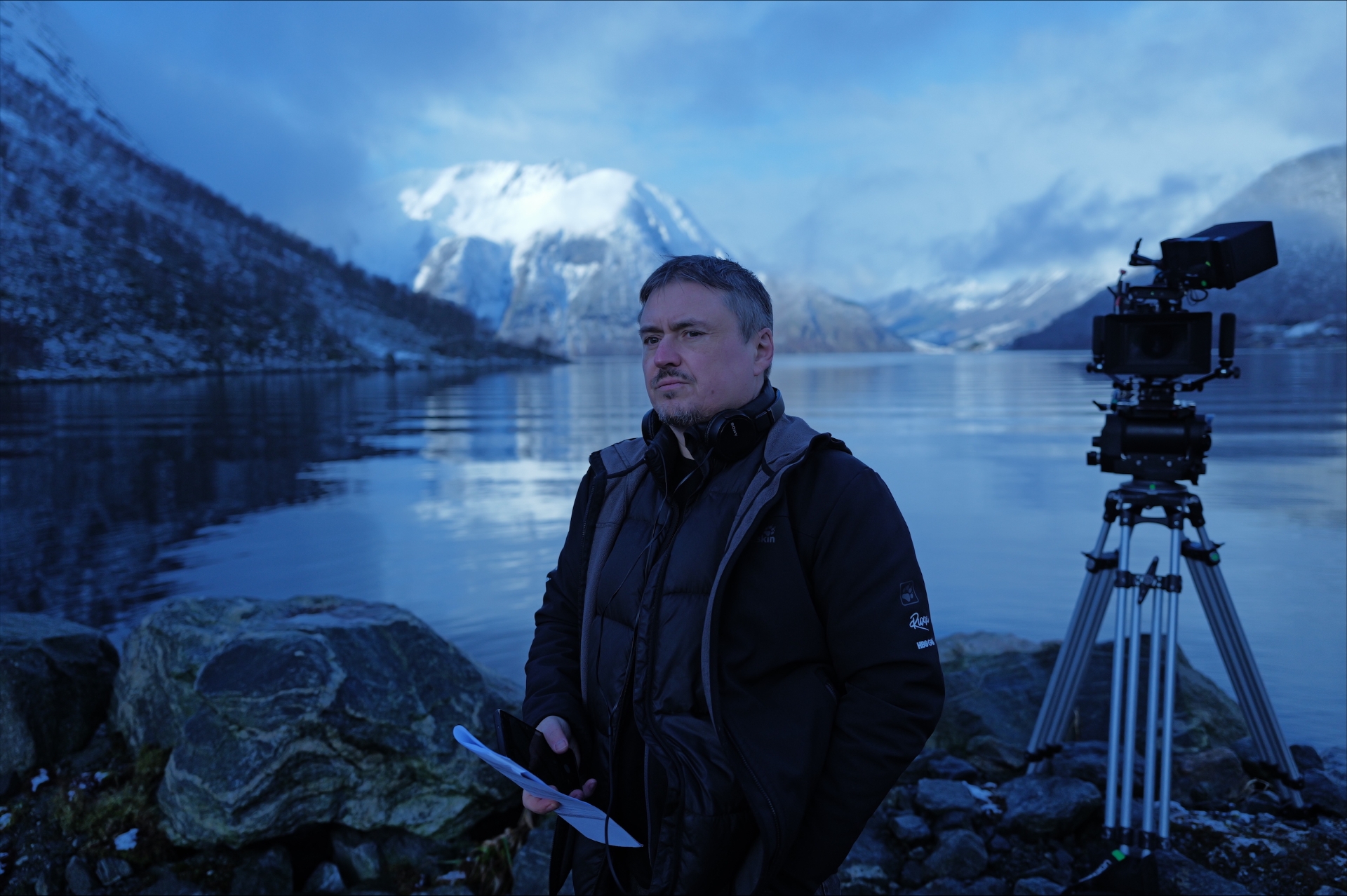foto. mann i halvfigur står foran fjordlandskap i blåtoner med kamera på stativ bak seg