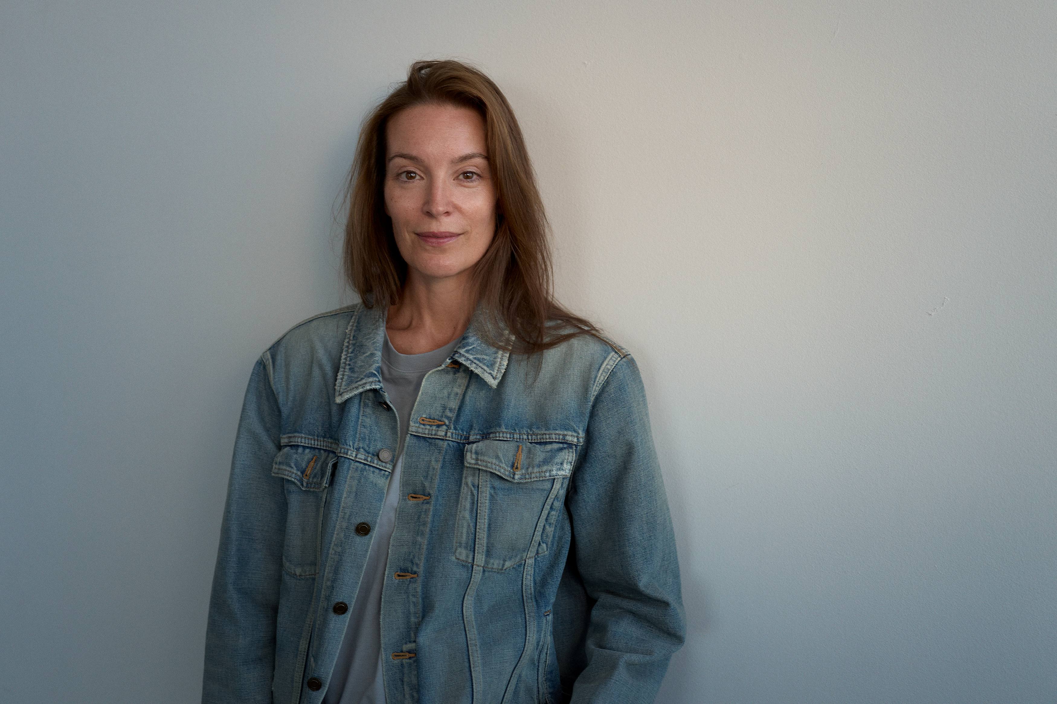 foto. kvinne med lys denimjakke og langt hår ser mot kamera, portrett halvfigur innendørs