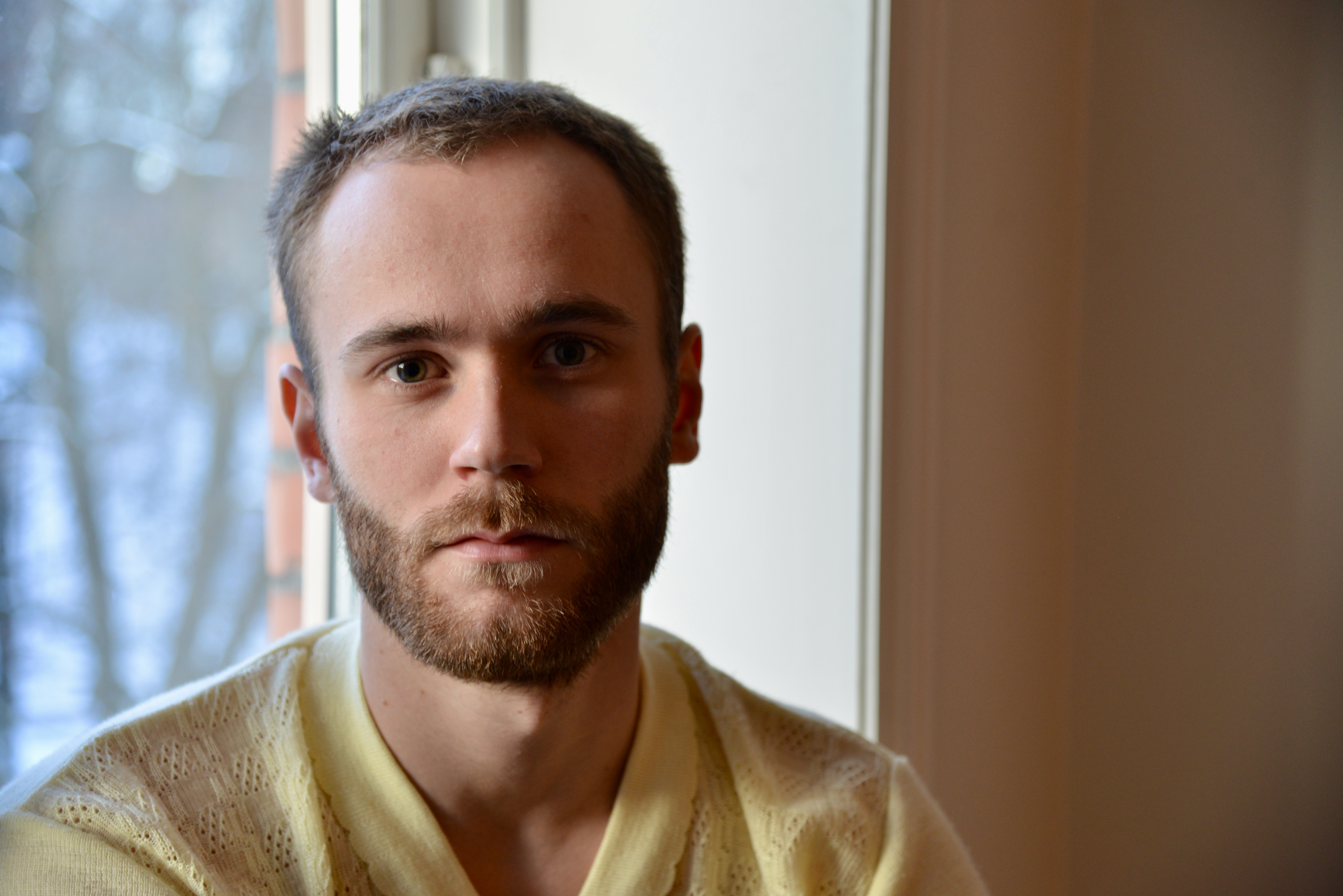 foto. portrett av ung mann med kort hår og skjegg ved et vindu som ser inn i kamera