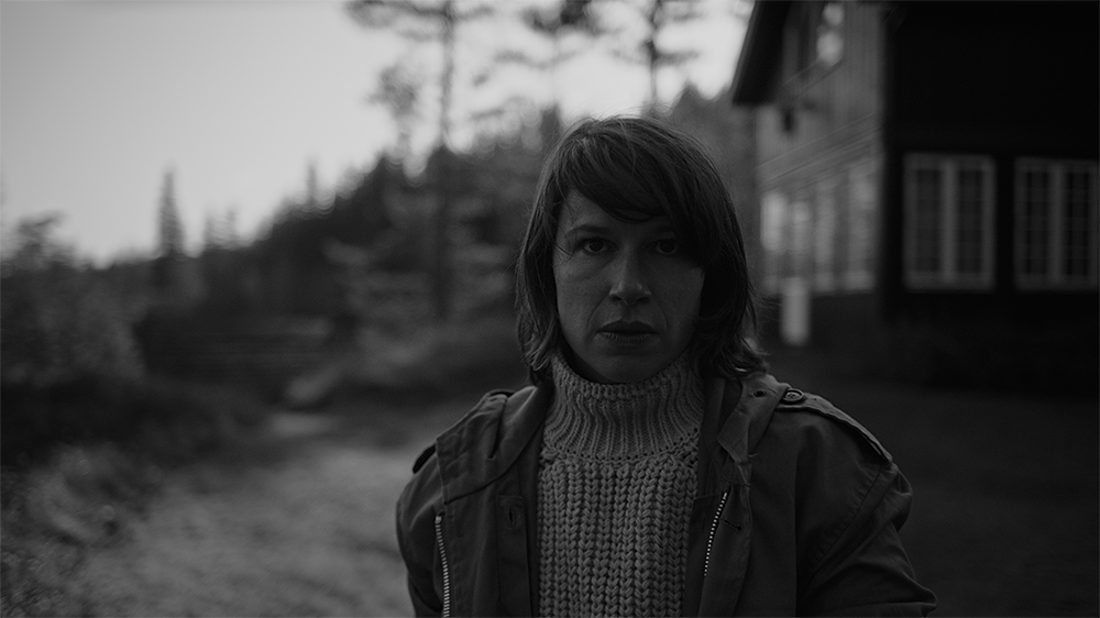 Foto: Sort hvitt portrett av en kvinne med strikkegenser og jakke. I bakgrunnen ser man et hus og skog.
