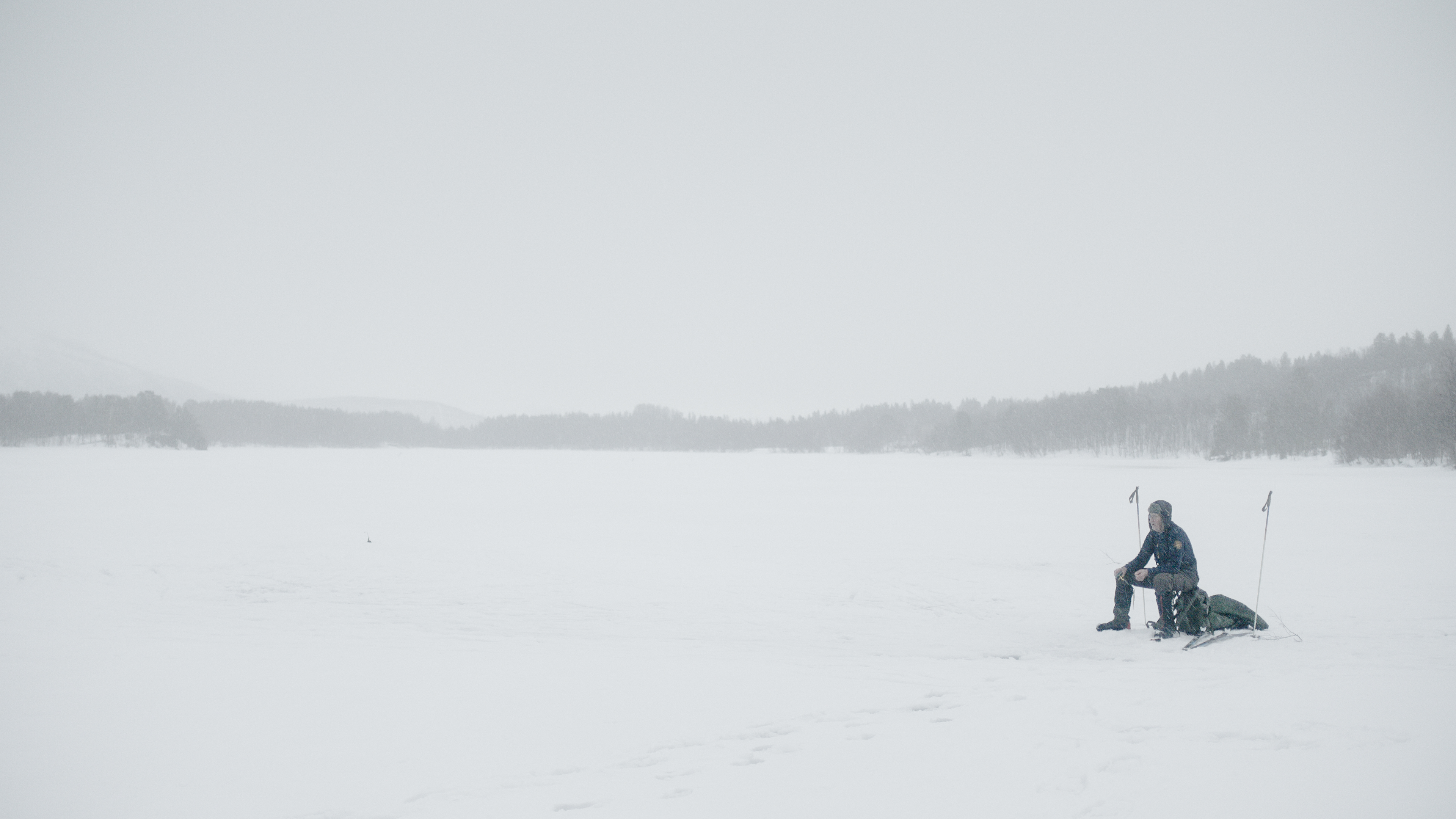 foto. person i svart sitter på en ryggsekk mellom to skistaver på en snødekt slette, bilde tatt på lang avstand
