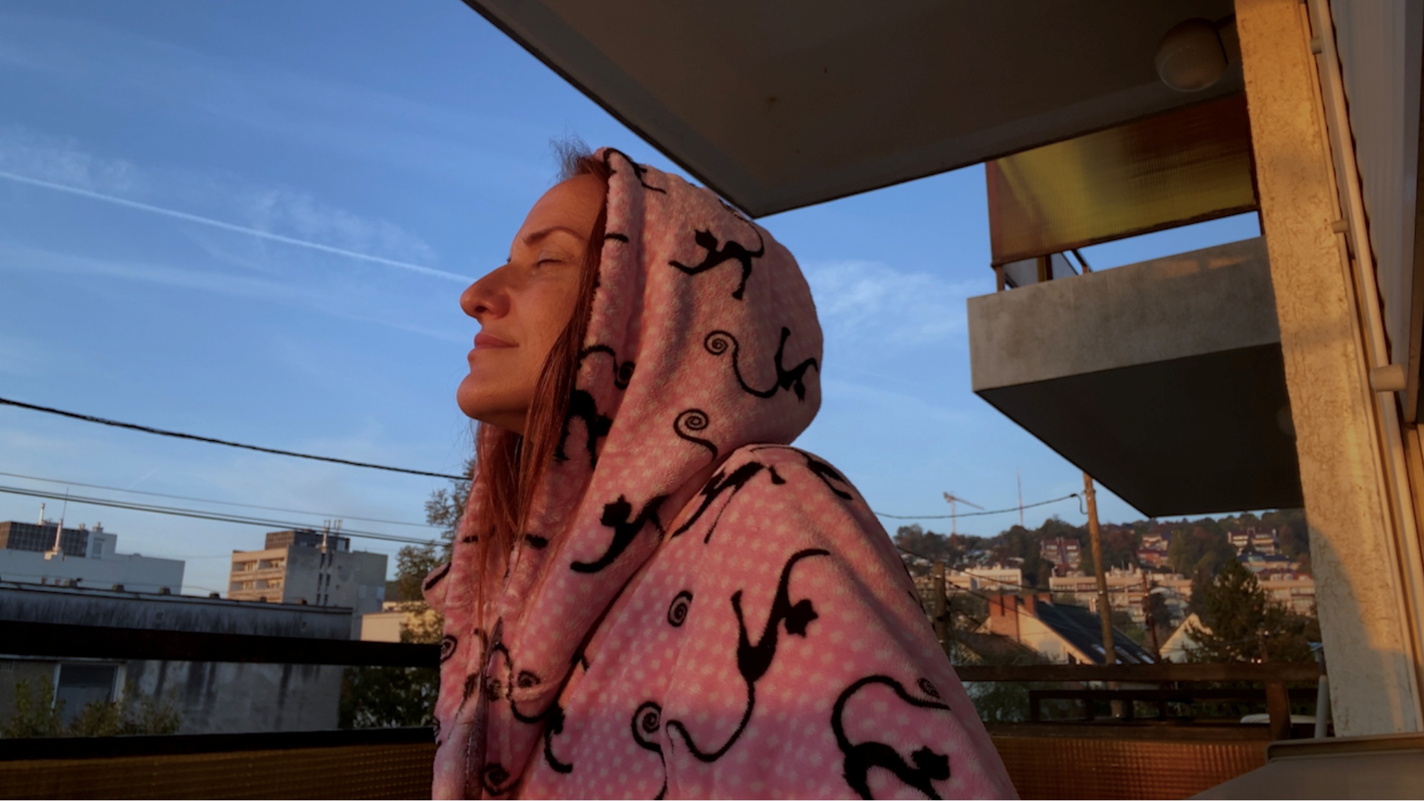 foto. kvinne med hettejakke i profil halvfigur myser mot sola på en balkong med boligblokker i bakgrunnen