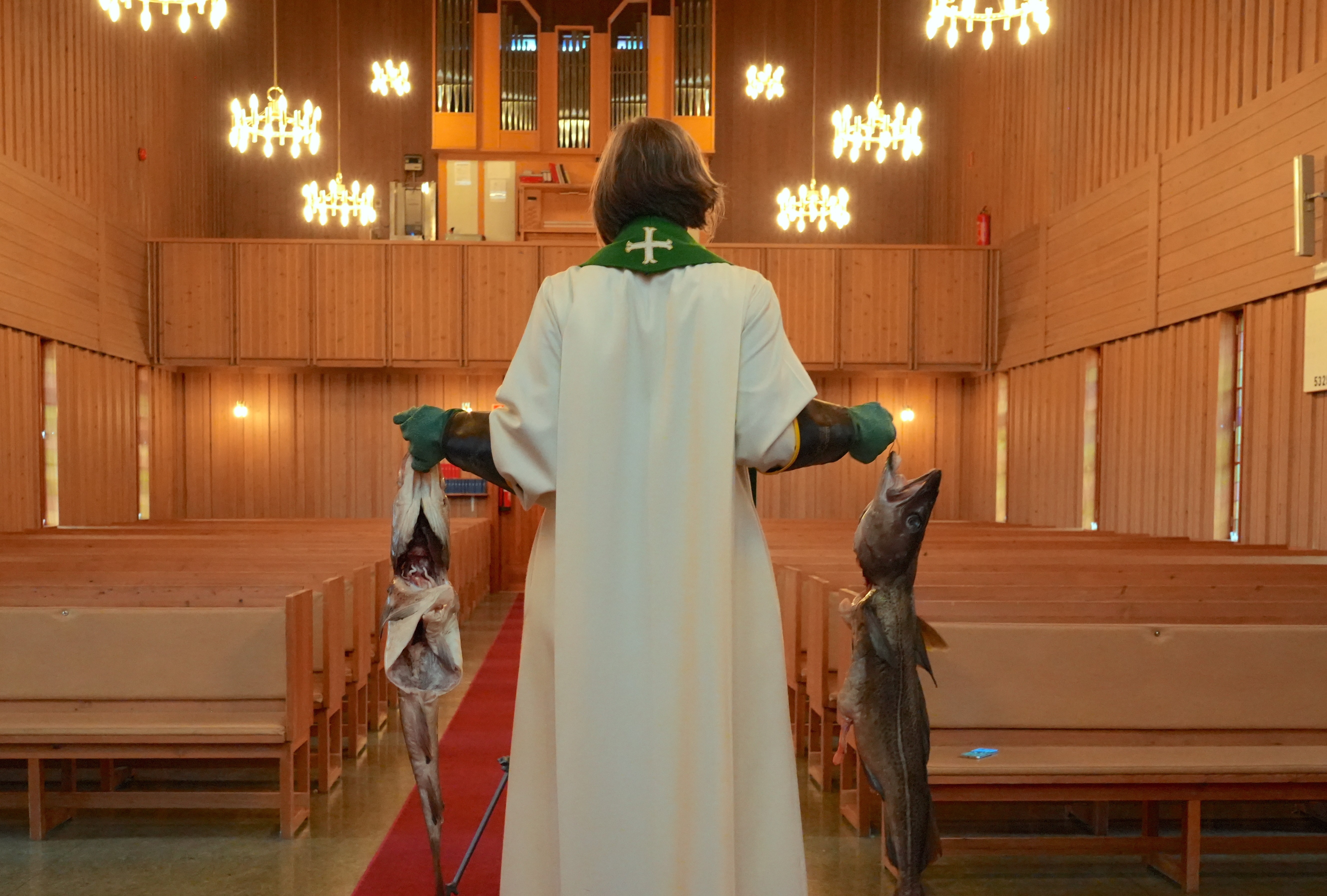 foto. person i prestedrakt bakfra står i kirke med en stor fisk i hver hånd