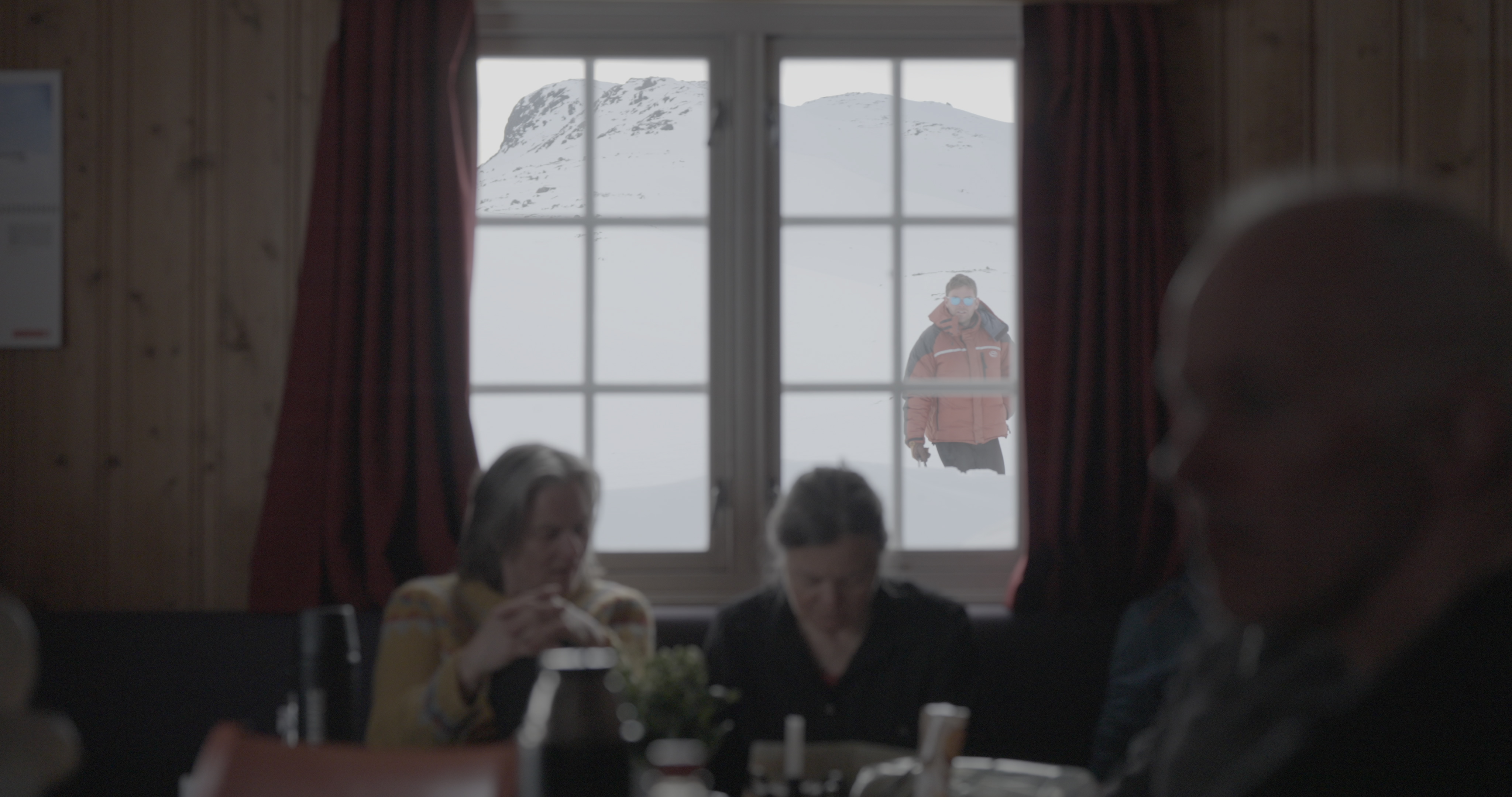 foto. tre personer rundt et bord inne i en hytte med en mann som ser inn utenfra i snøen