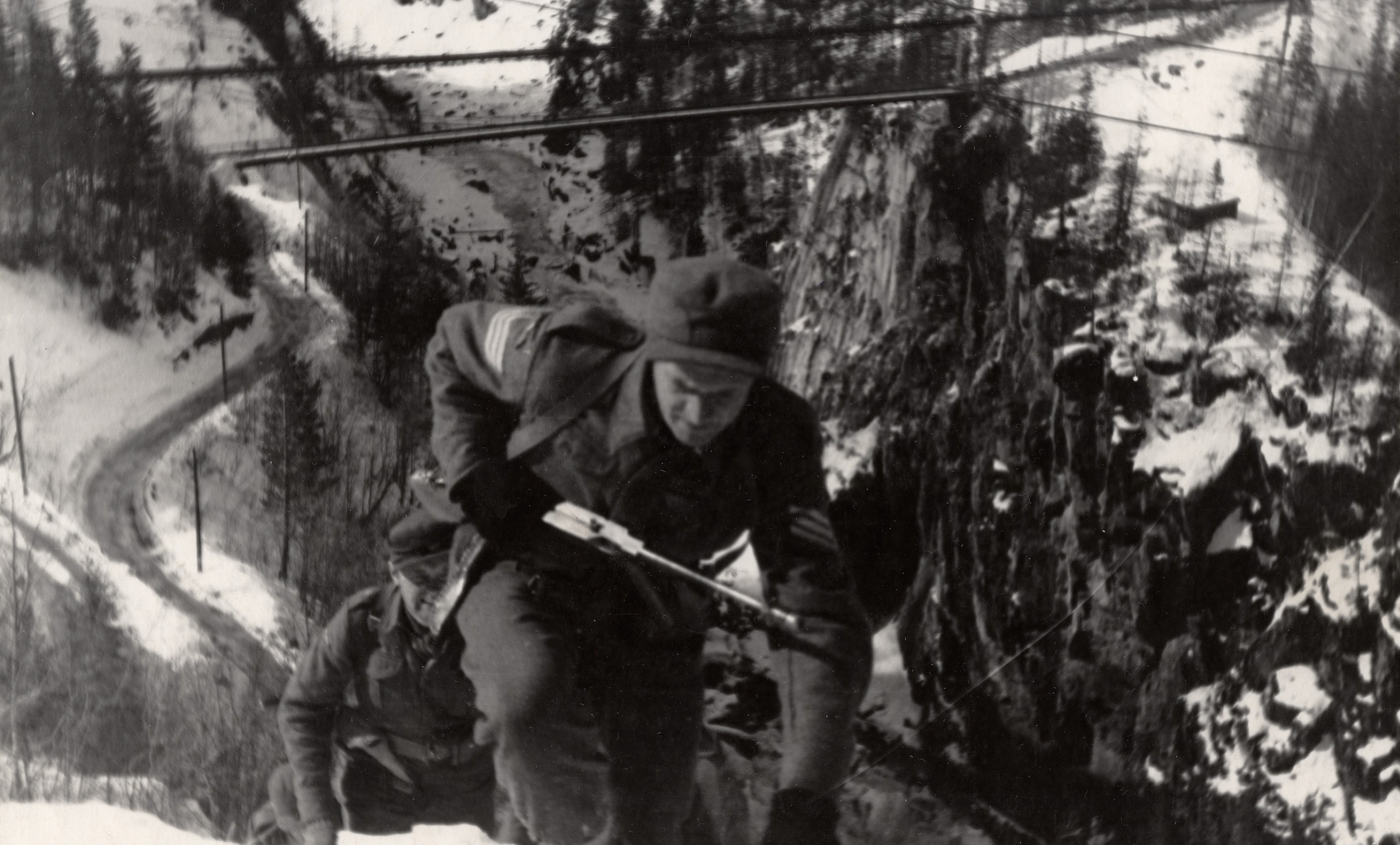 foto. Svart-hvitt-bilde av to menn i militæruniformer og med våpen i hånd som klatrer opp et bratt juv.