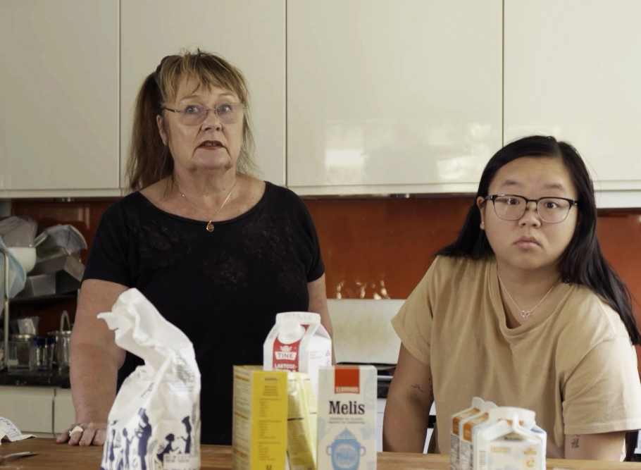 foto. ung kvinne foran og eldre kvinne bak står ved kjøkkenbenk med melpose, meliskartong, melk og andre bakeingredienser