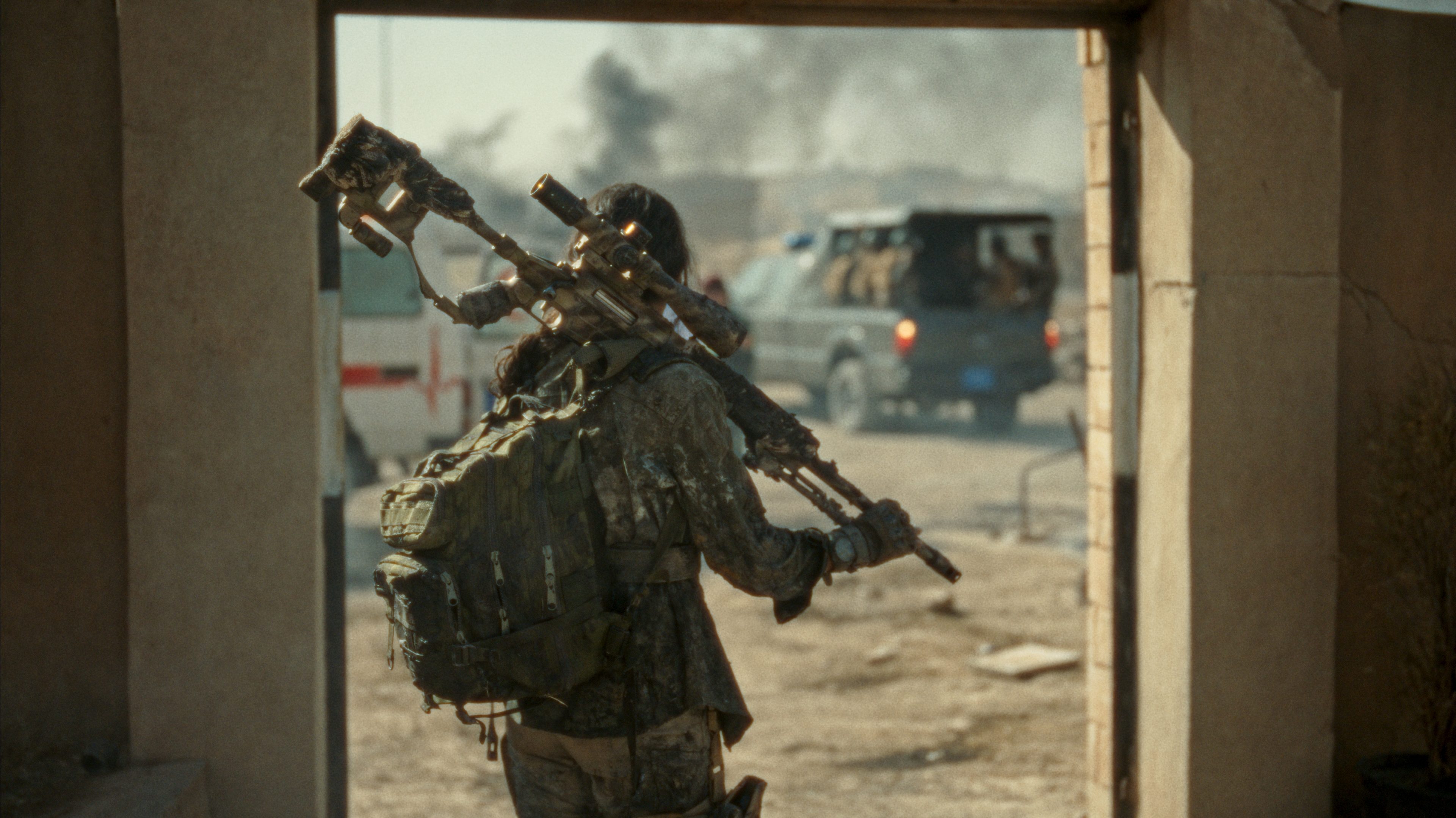 Photo. Person with big sniper rifle stands in doorway with cars in the background.