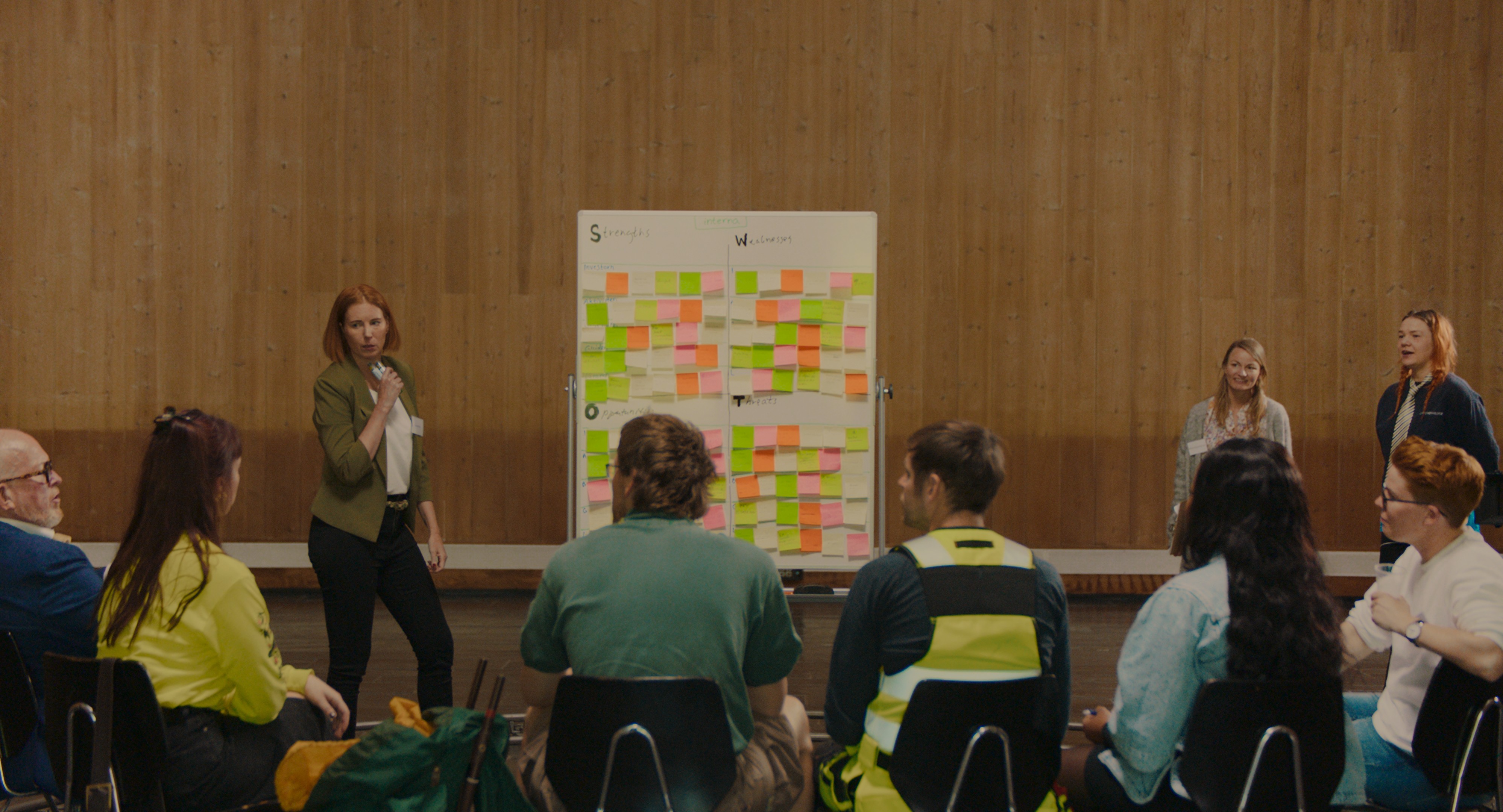 foto. en kvinne står og snakker i en mikrofon foran en tavle full av fargerike post it-lapper i en sal med åtte andre personer