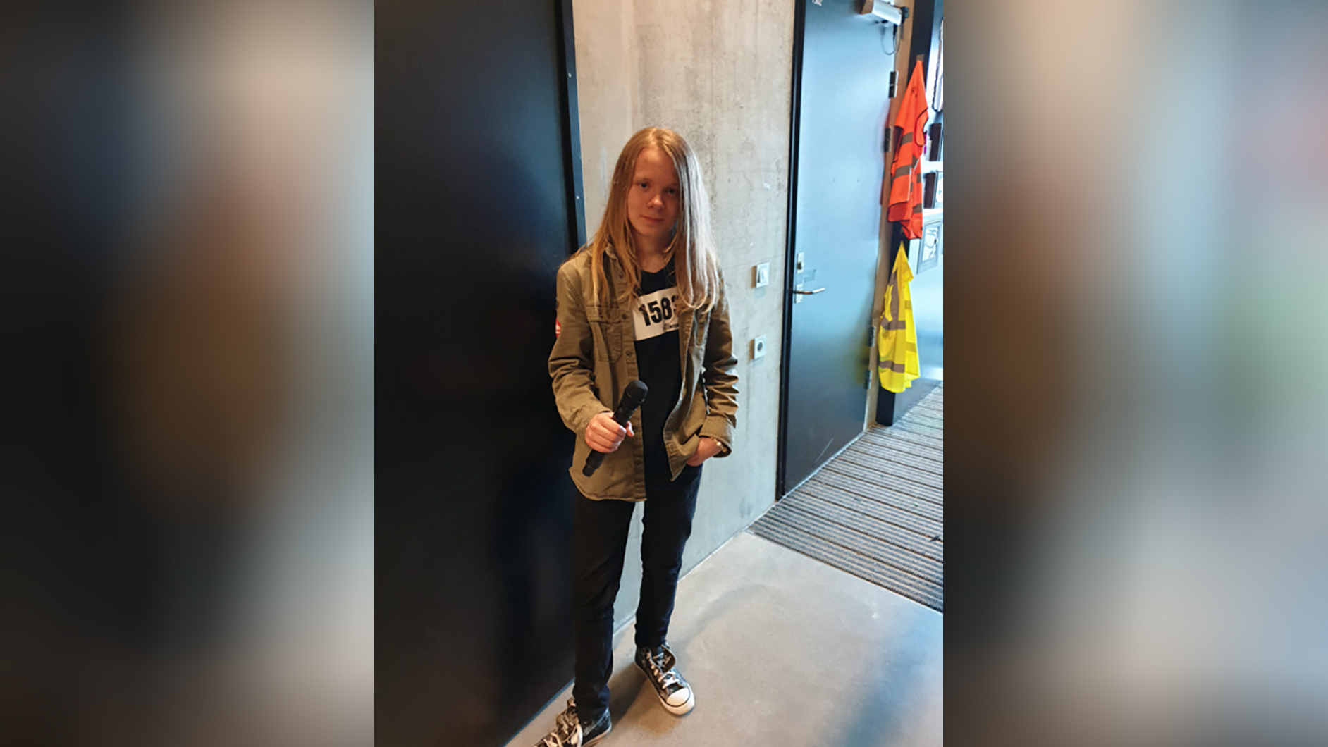 bilde av ung person med langt hår som står foran en dør med en mikrofon i hånden og ser inn i kamera