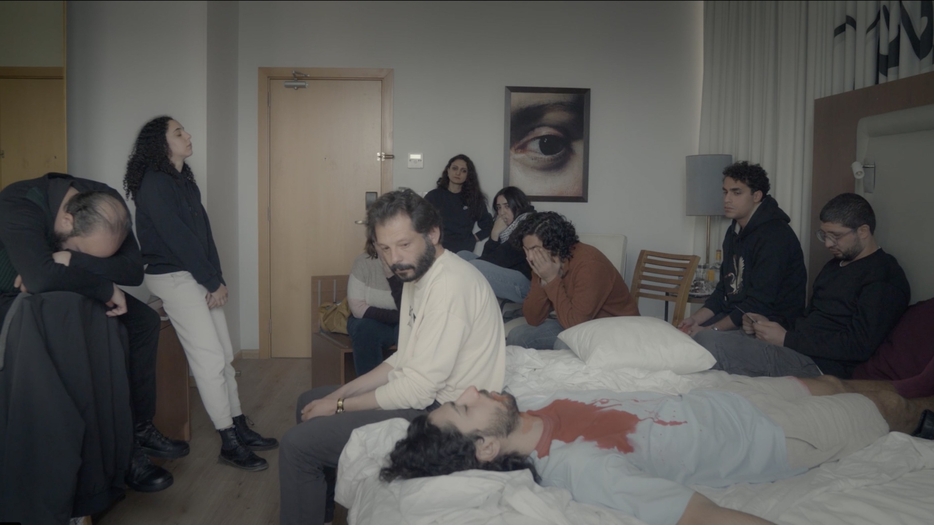 Foto. Menn og kvinner sitter gråtende rundt person som ligger med lukkede øyne med rød farge - noe som skal forestille blod - på brystet. Det er en iscenesettelse, altså ikke en reell situasjon.