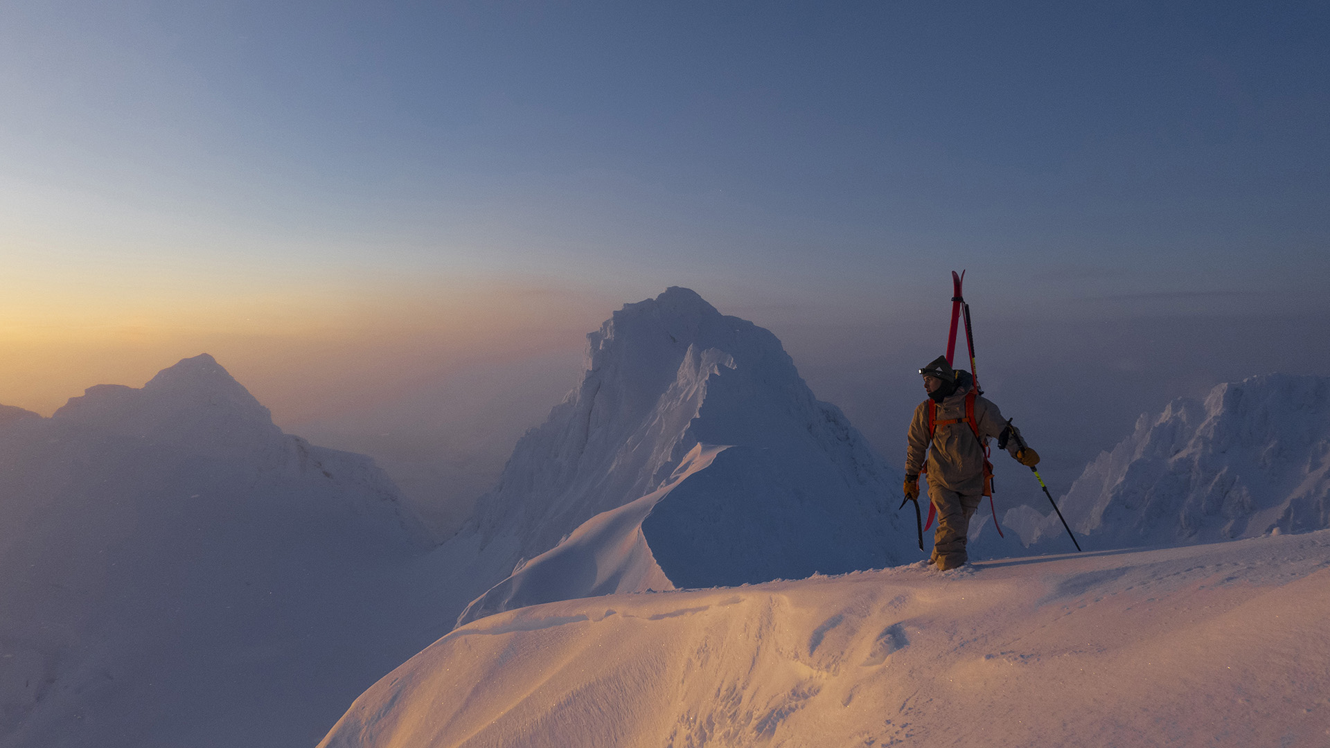 Foto: En mann går på toppen av snøkledd fjell med ski på ryggen ved soloppgang.