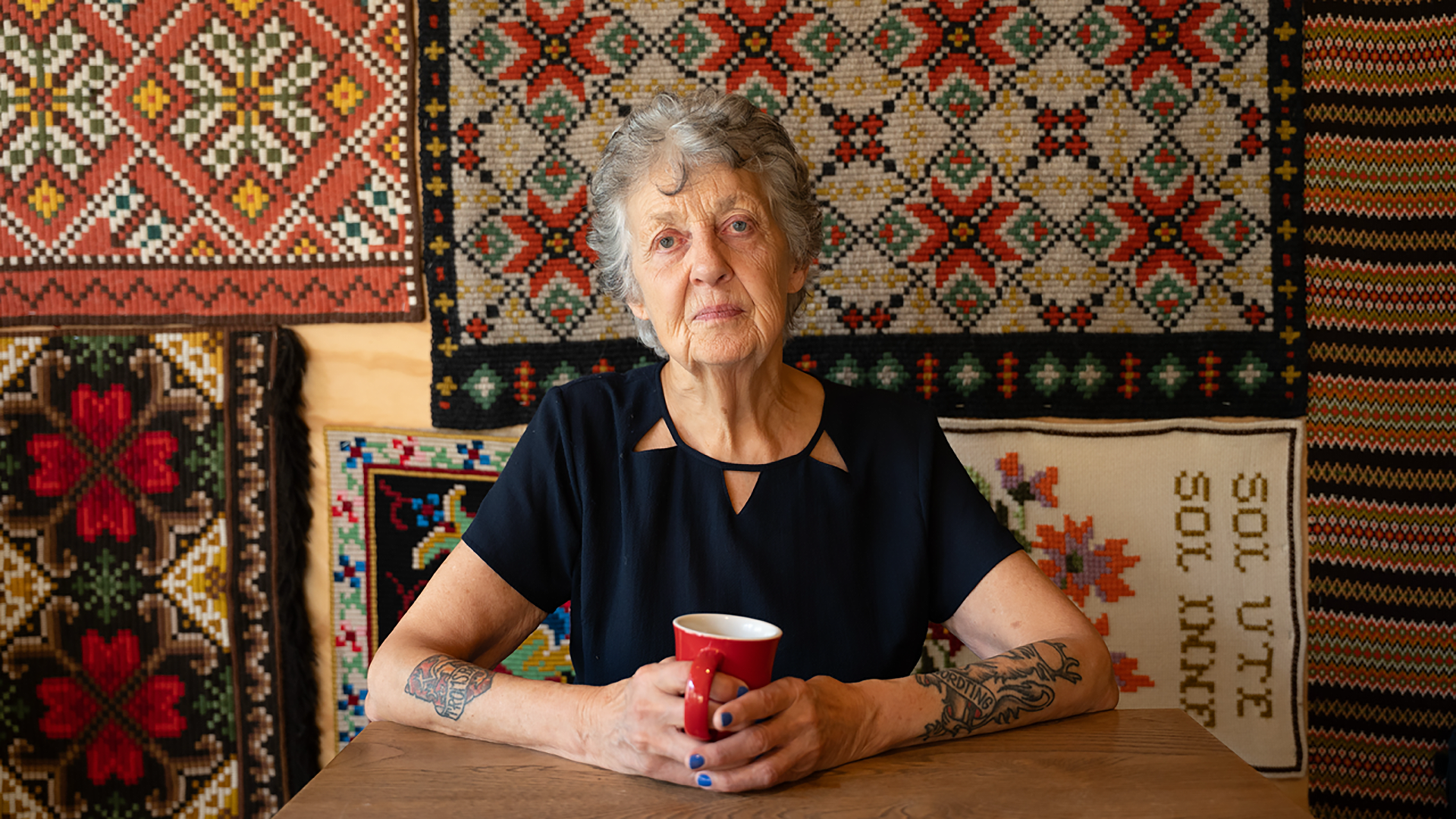 Foto. Portrett av en eldre kvinne. Hun holder i en kopp, og på veggen bak henne henger det flere broderte veggtepper