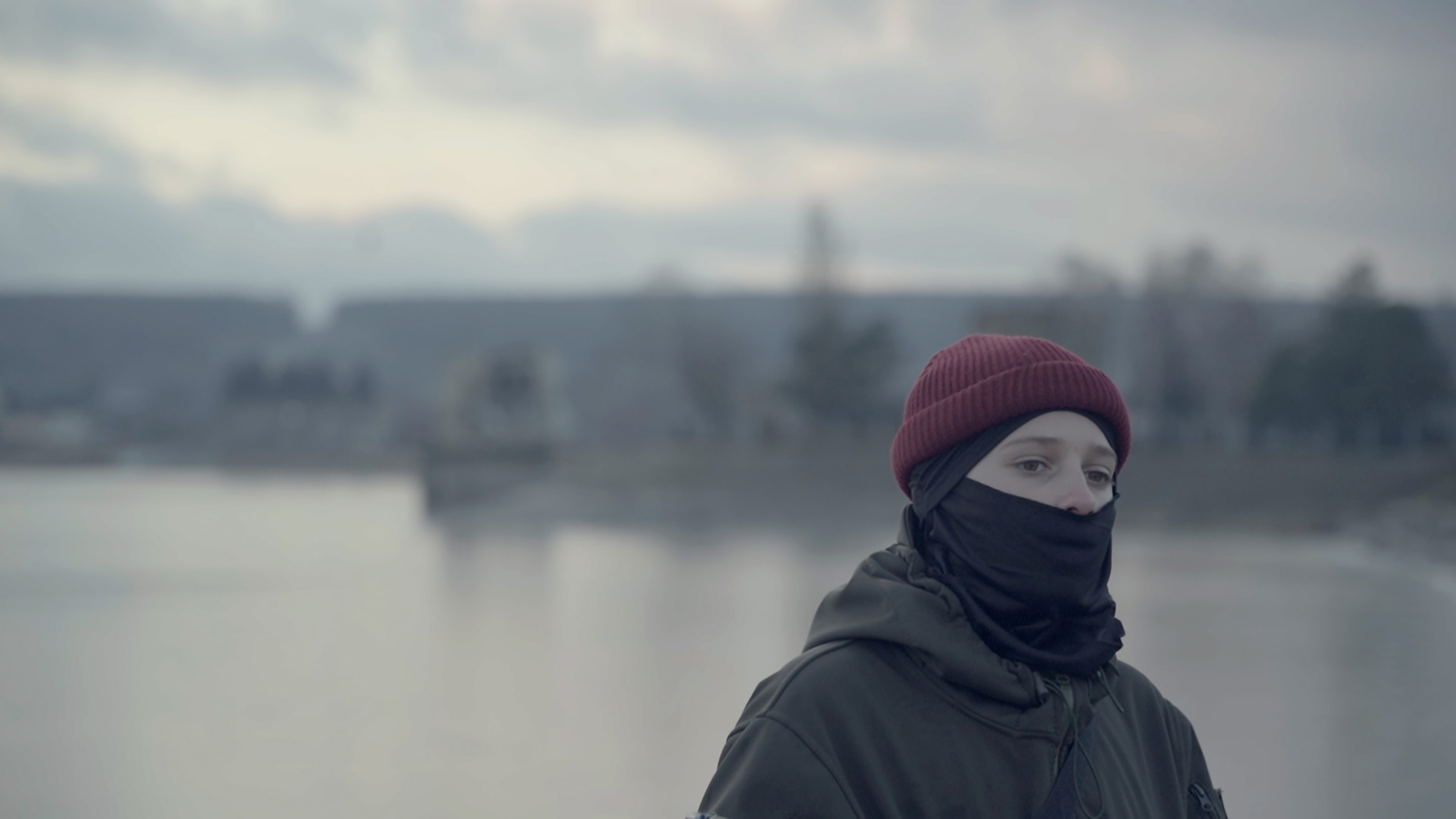 foto. person med rød lue og svart skjerf foran munnen står ved en elv med bygninger i bakgrunnen