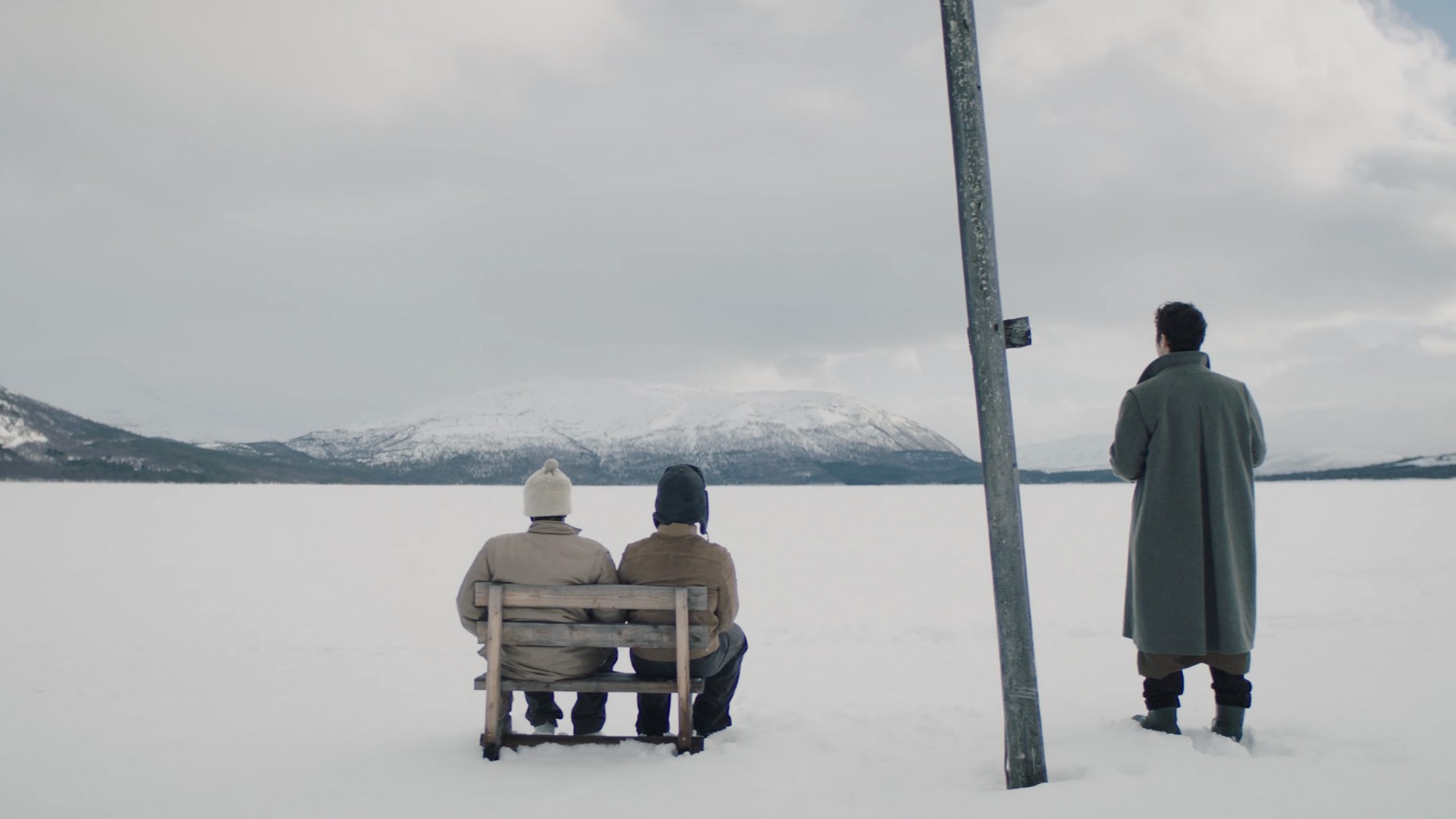 Foto. Tre menn er på en snøvidde og ser mot et fjell i bakgrunnen, to av de sitter på en benk mend den tredje står ved siden av.