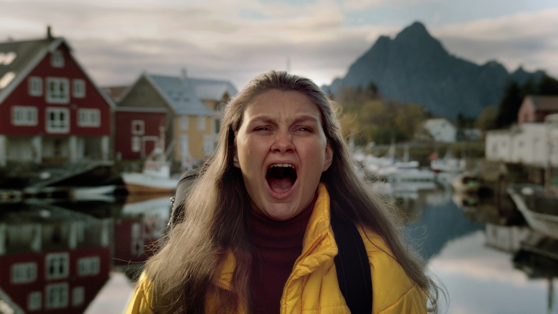 foto. kvinne i gul boblejakke står nærme kameraet og roper. i bakgrunnen ser vi stille vann, røde og hvite hus og et fjell.