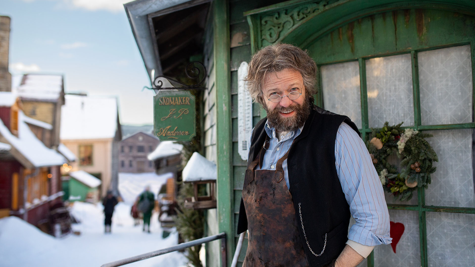 Foto. En med blå skjorte, mørk vest og skinnforkle står foran et grønt trehus og smiler mot kamera.