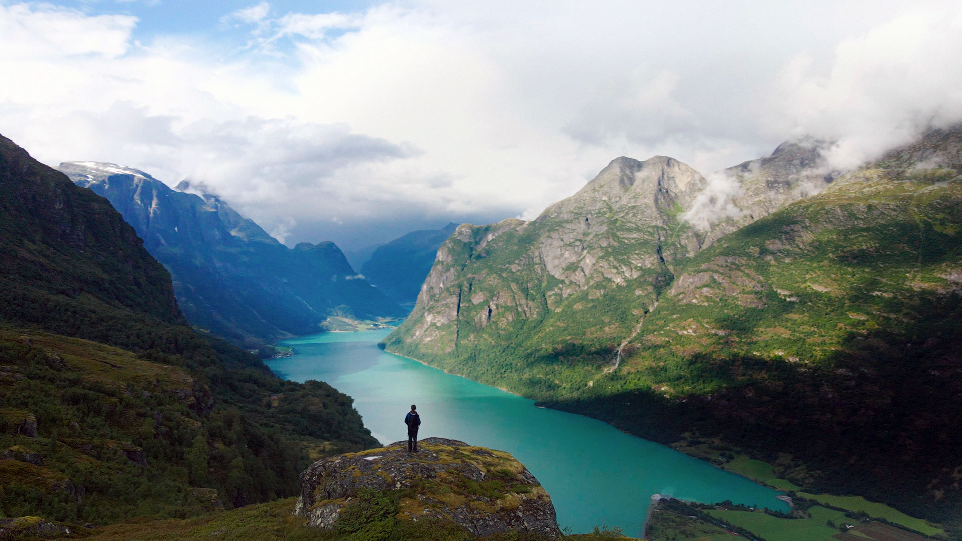 foto. en person står på en klippe og ser utover en stor fjord og fjellene rundt.