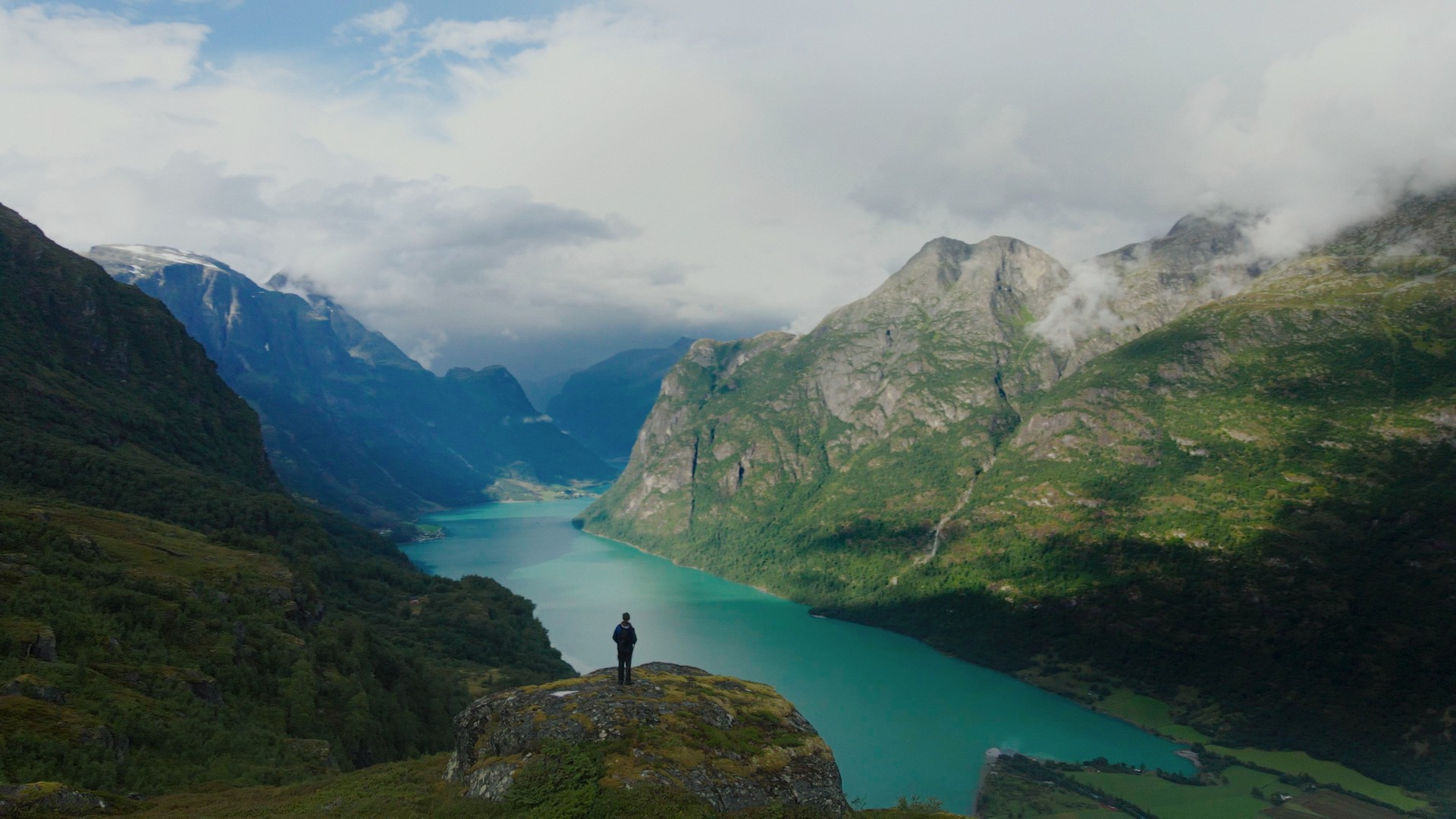 Foto. En person står på en liten fjelltopp foran et stort fjellandskap og en lang fjord.