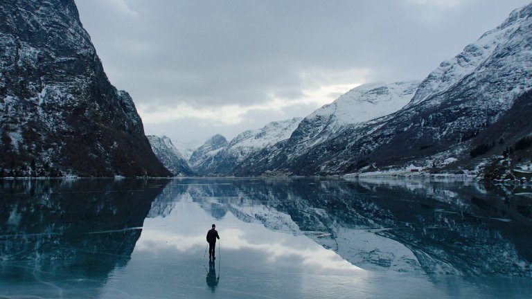 foto. en mann går alene på isen, omringet av fjell som er reflektert i isen han går på.