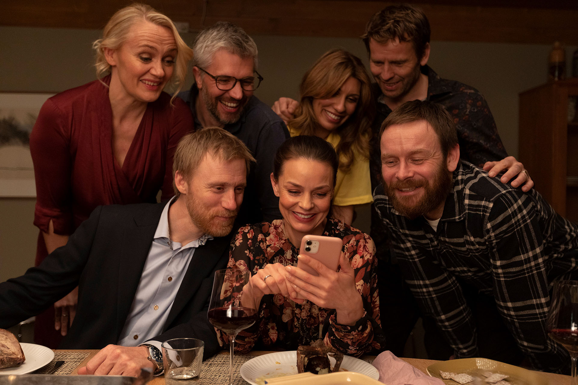 Foto. En kvinne sitter ved et bord og sveiper på en telefon, seks andre personer står rundt henne og ser på telefonen.