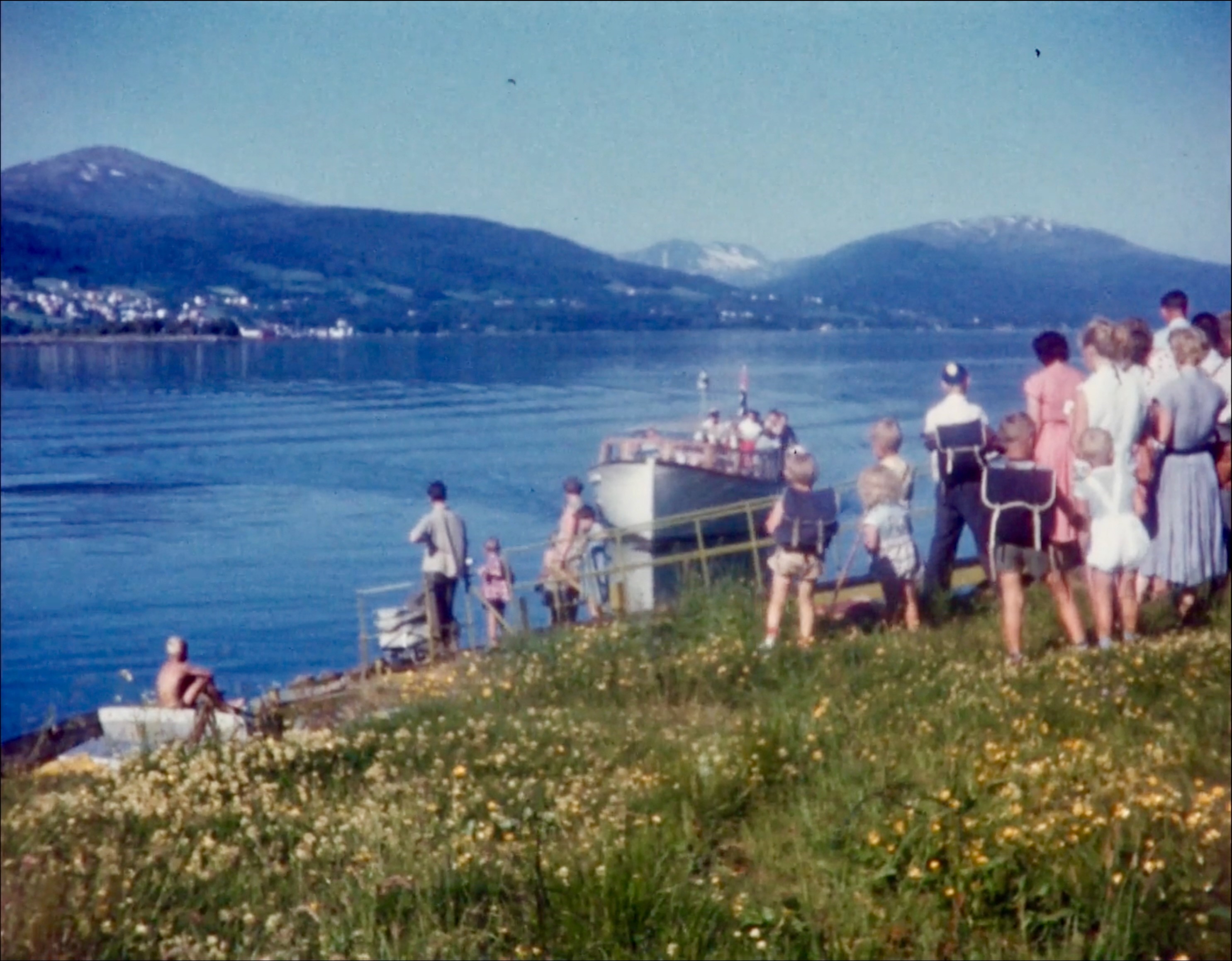 Foto. En gruppe mennesker står ved vannkanten og venter på en båt som som er like ved strandkanten.