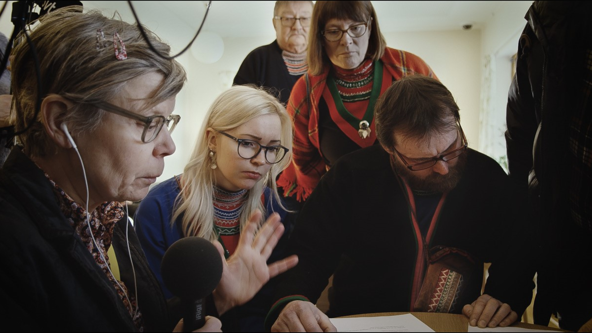 Foto. Fem personer står rundt en pult, kvinnen i forgrunnen har øretelefoner i ørene og gestikulerer med sin venstre hånd.