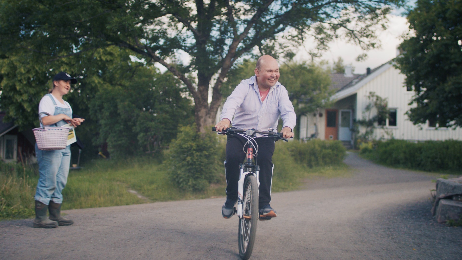 Foto. En mann sykler smilende nedover en vei, til venstre i bildet står det en kvinne som holder en kurv.