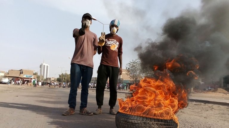 foto. to menn med hvite masker på står bak et bildekk som brenner.