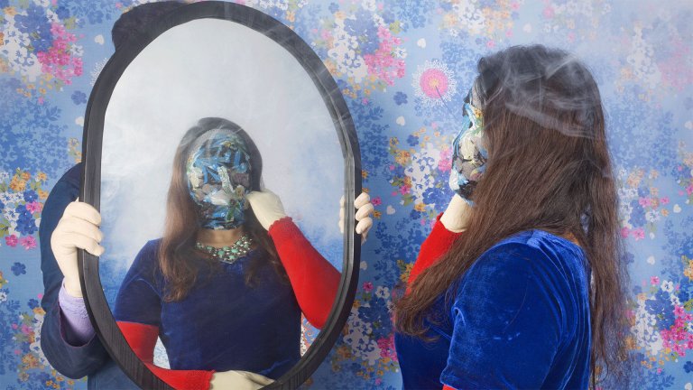 foto. en kvinne står foran et speil, hun har på seg en maske som dekker hele ansiktet med blomstrete mønster. bak speilet er det en vegg med blomstrete mønster.
