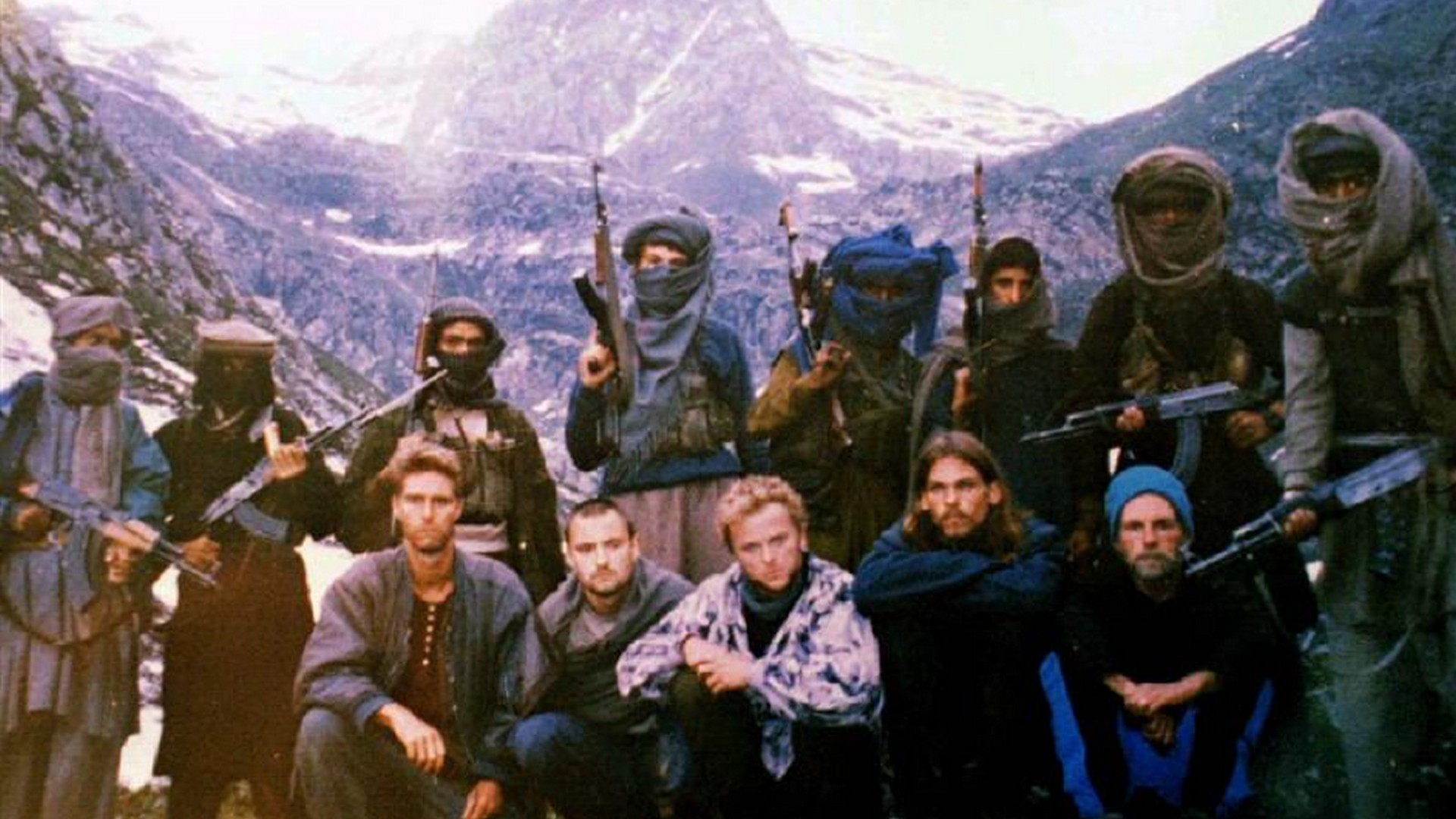 Foto. En gruppe mennesker står foran et fjellanskap, flere i gruppen holder automatvåpen.