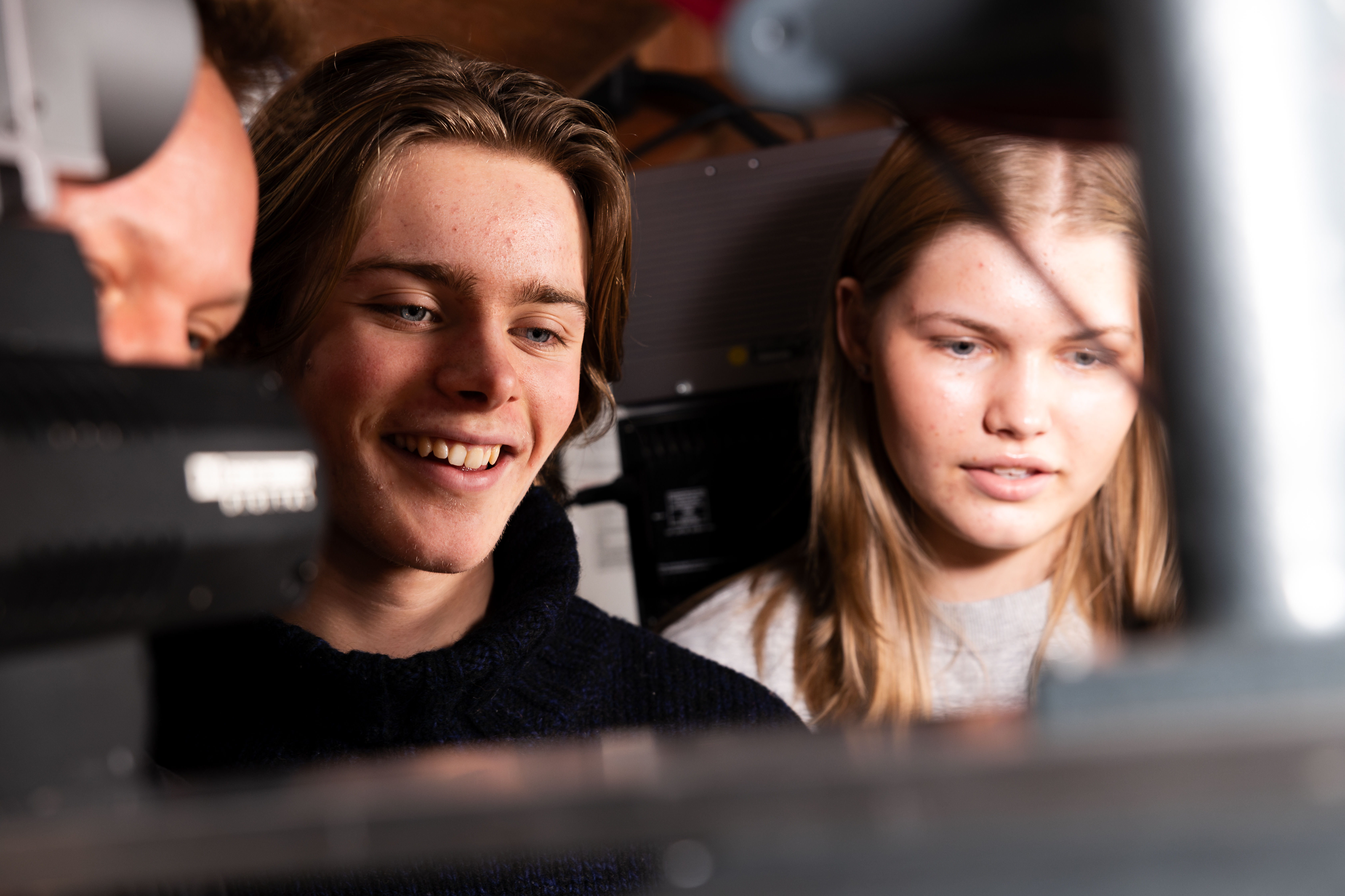 Foto. Ung gutt og jente smiler og ser på en skjerm. Man skimter også pannen og øynene til enda en person.