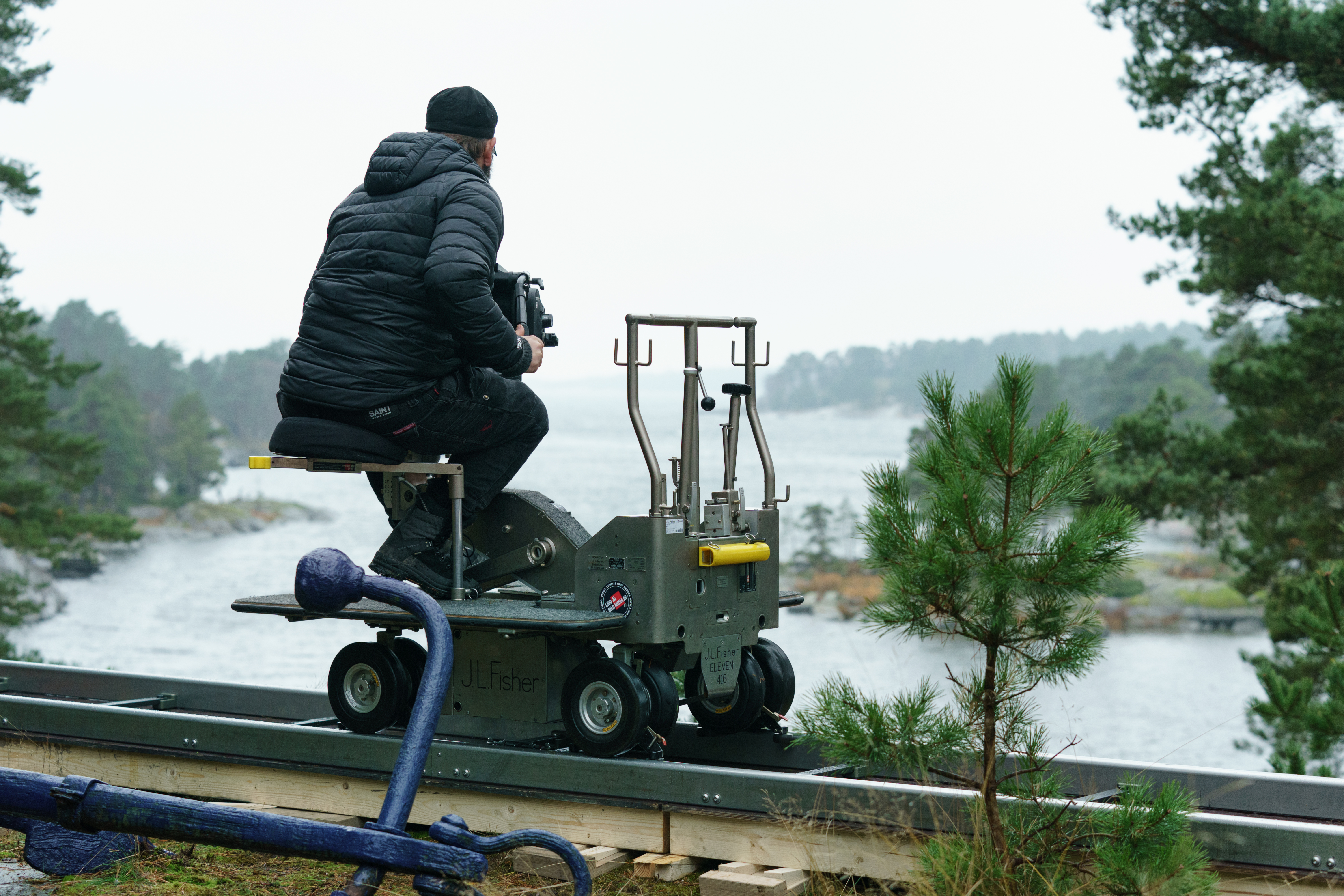 Foto.Kamermann sitter på liten vogn og filmer med ryggen til, vi ser ut over en fjord.