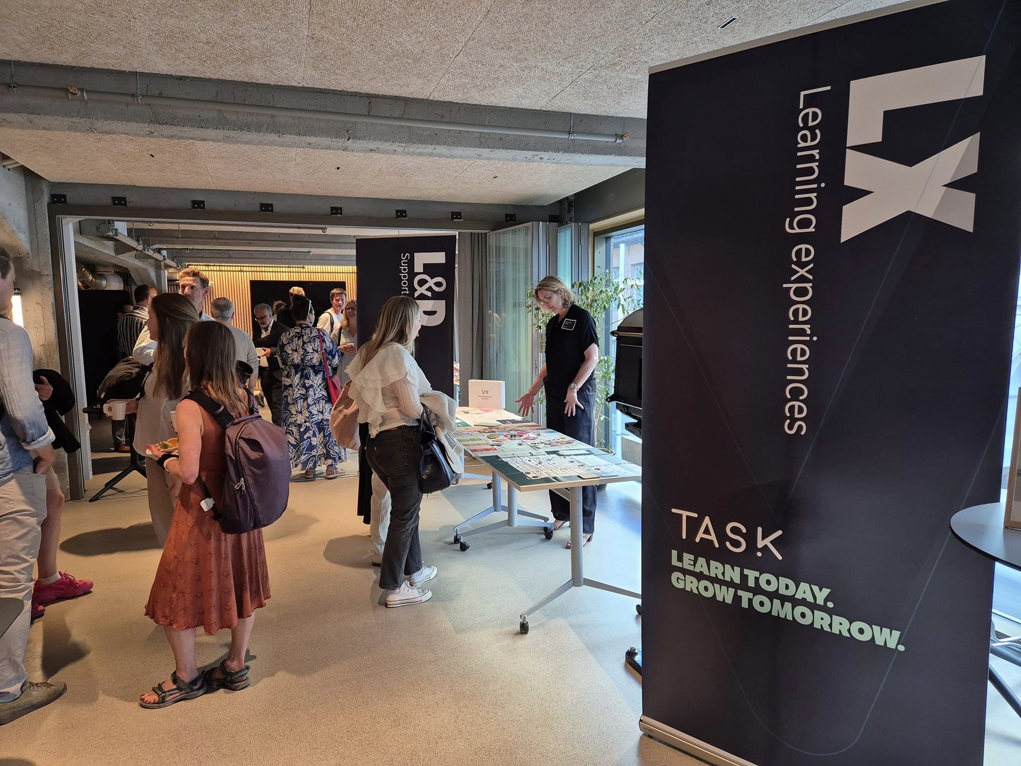 Foto:. Flere mennesker på seminar som står rundt bord og mingler rundt på gulvet. Flere Roll-ups med reklame for Task Learning Experience.