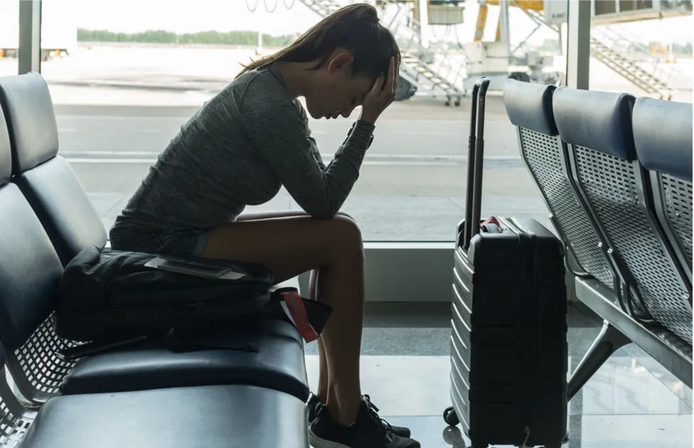 female passenger with carry-on at airline terminal seat, stressed, head bowed