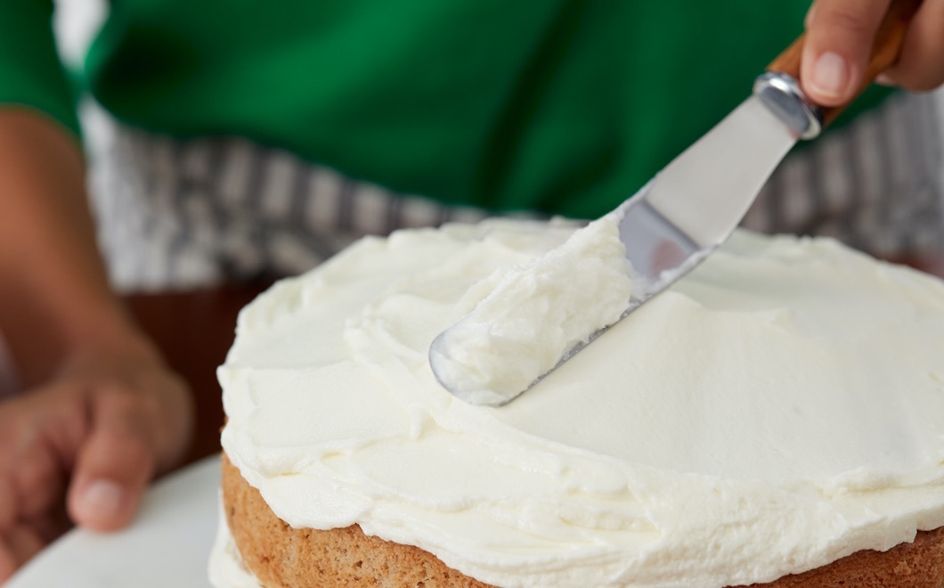 closeup of knife spreading creamy frosting on a two-layered cake