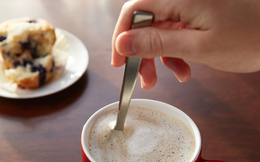 closeup of hand stirring a latte and a muffin on a plate in the background