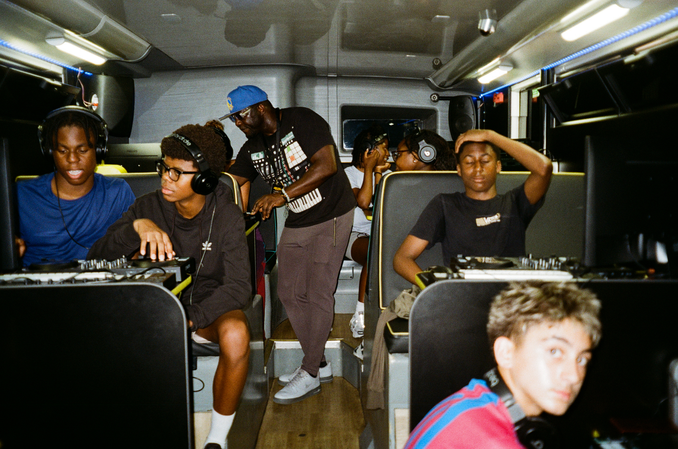 Group of people inside a bus or coach, some operating DJ equipment and turntables, with fluorescent lighting overhead.