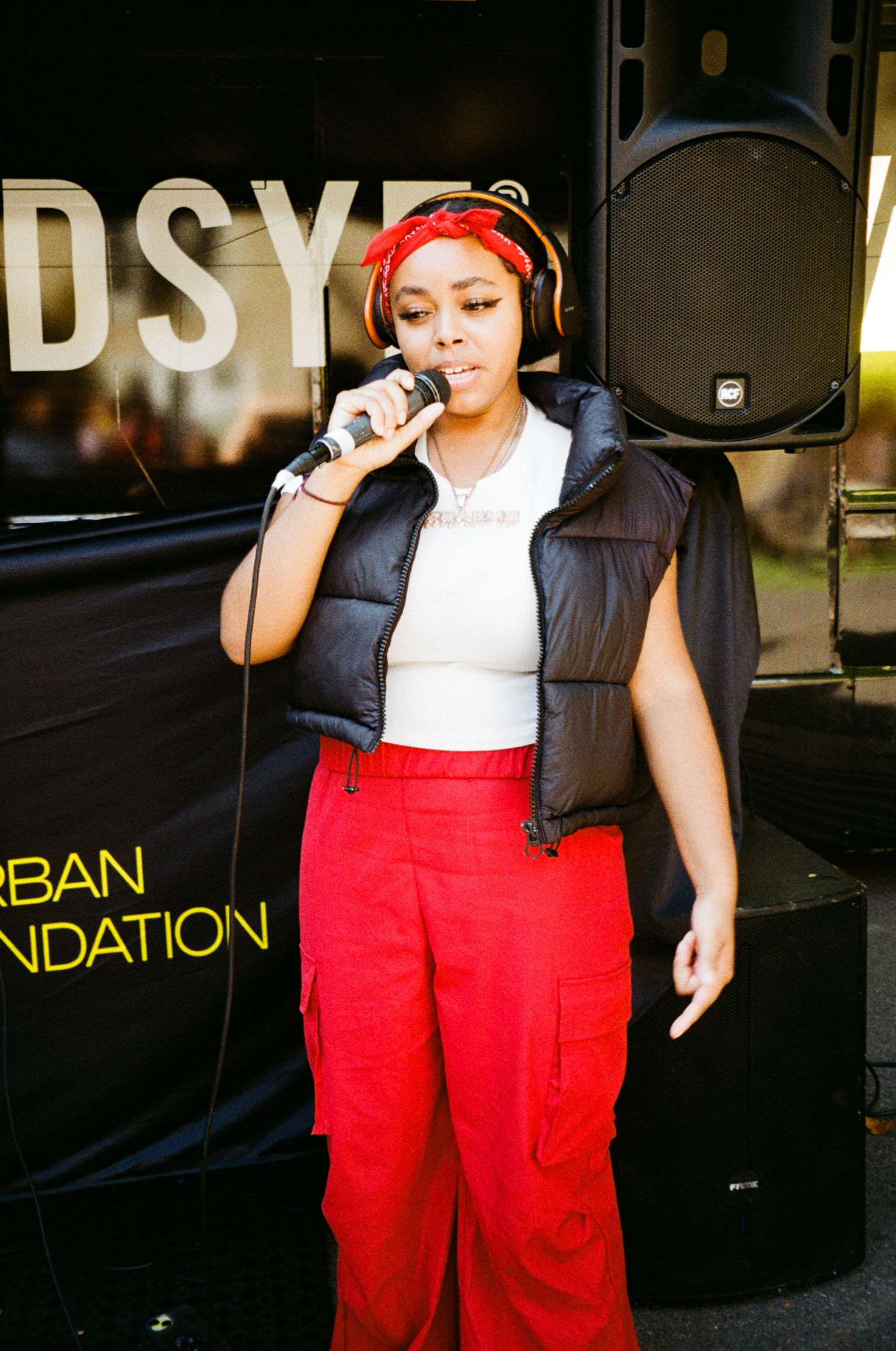 Woman in red headband and trousers, white top, black gilet speaking into microphone with large speakers behind her.
