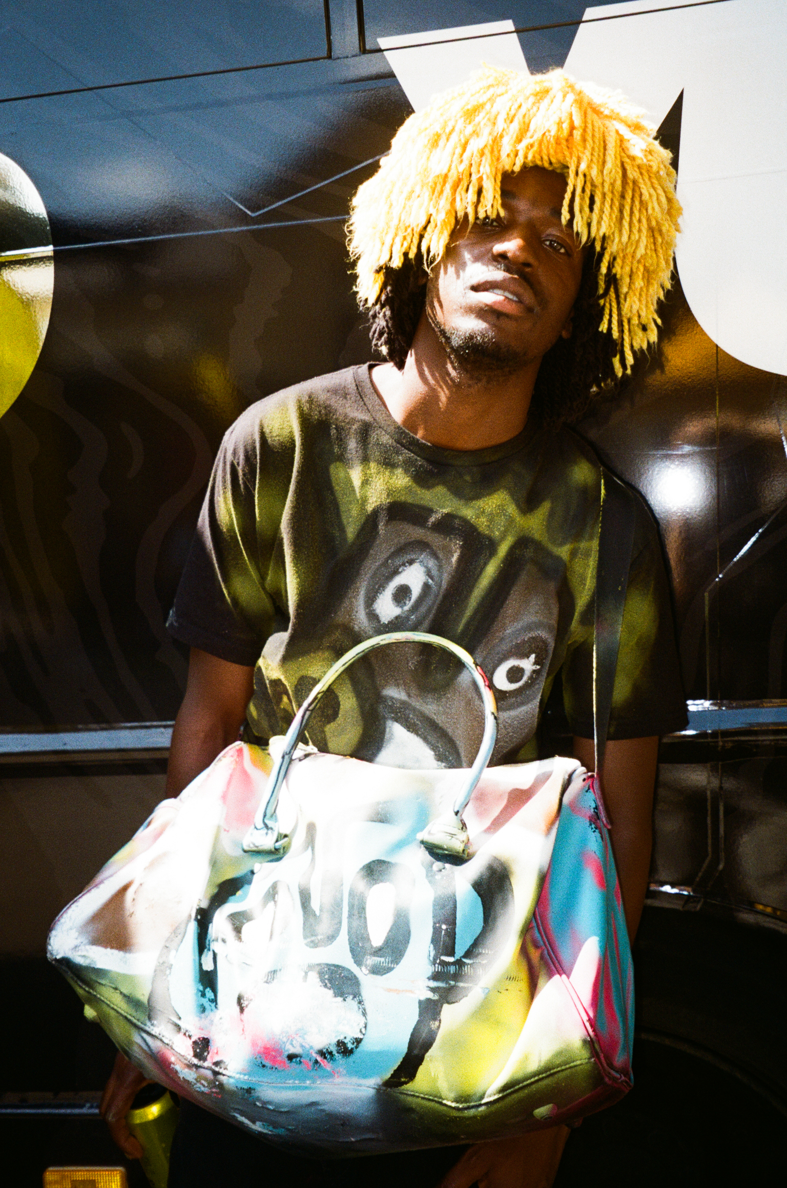Man wearing yellow fringe beanie and camouflage t-shirt holds multicoloured handbag with skull design, standing by grey vehicle.
