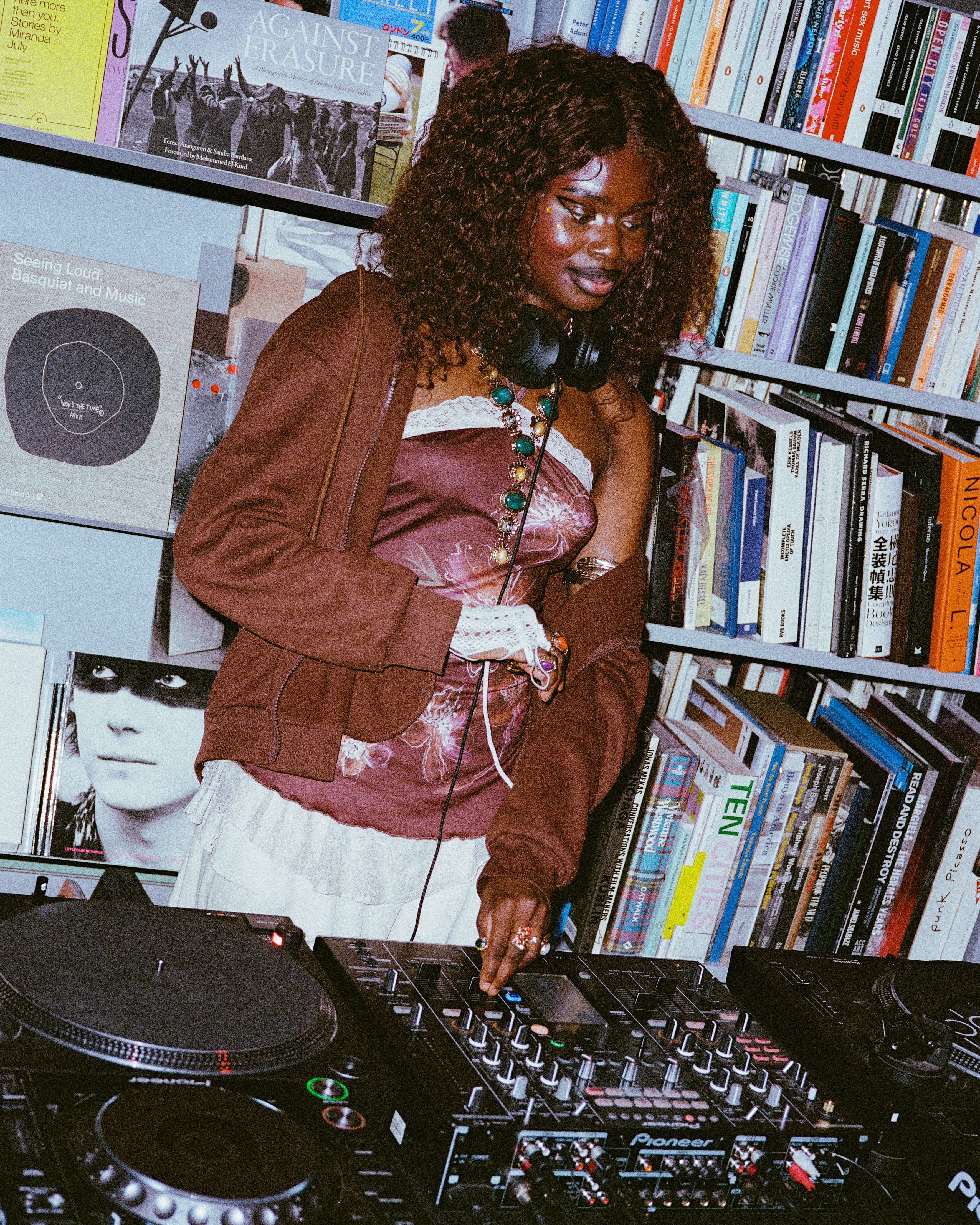 Woman with curly hair wearing headphones and brown cardigan, DJing with turntables and mixer in front of bookshelf filled with records and books.