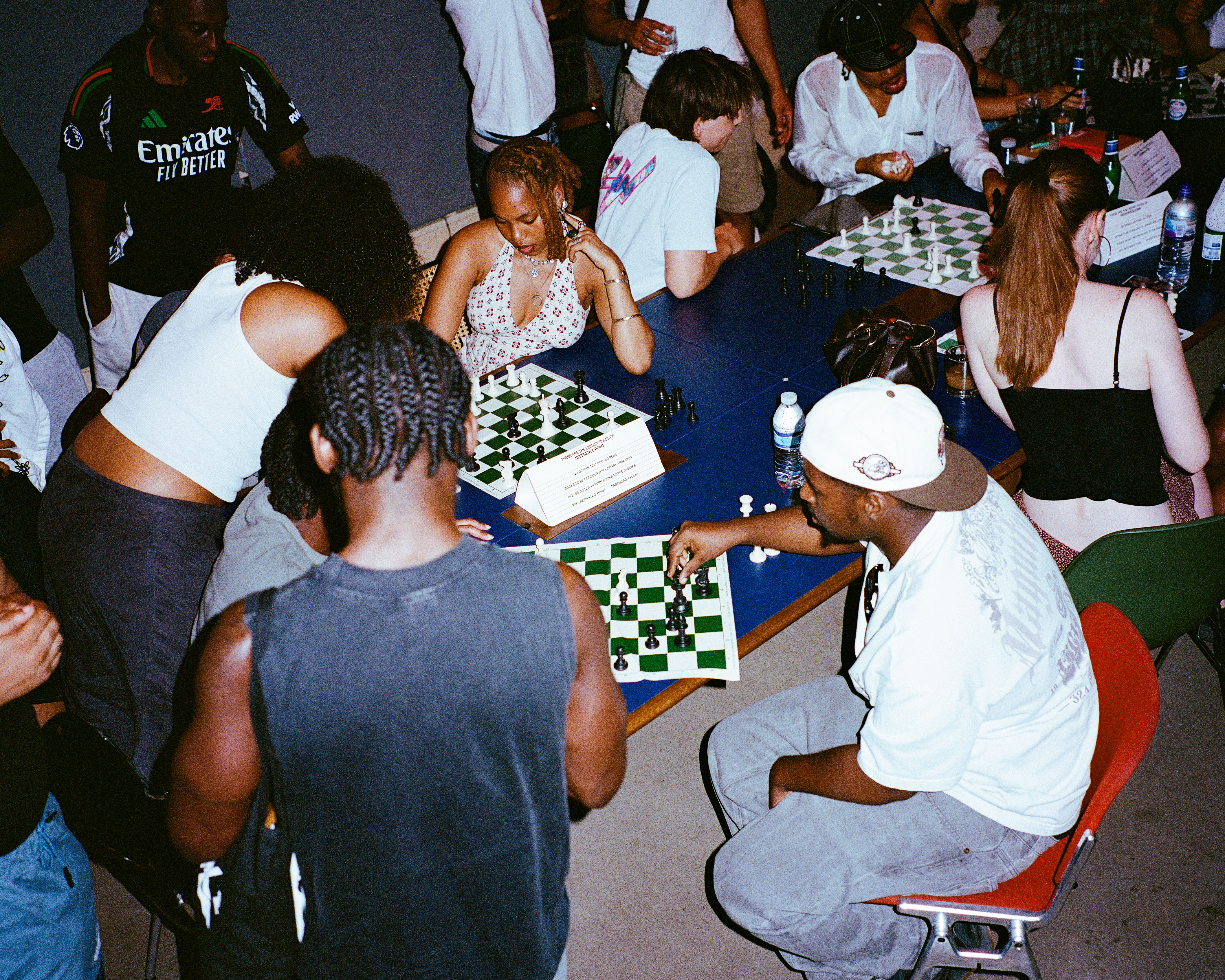 Group of people playing chess at outdoor tables, some standing and watching, casual clothing, evening lighting.
