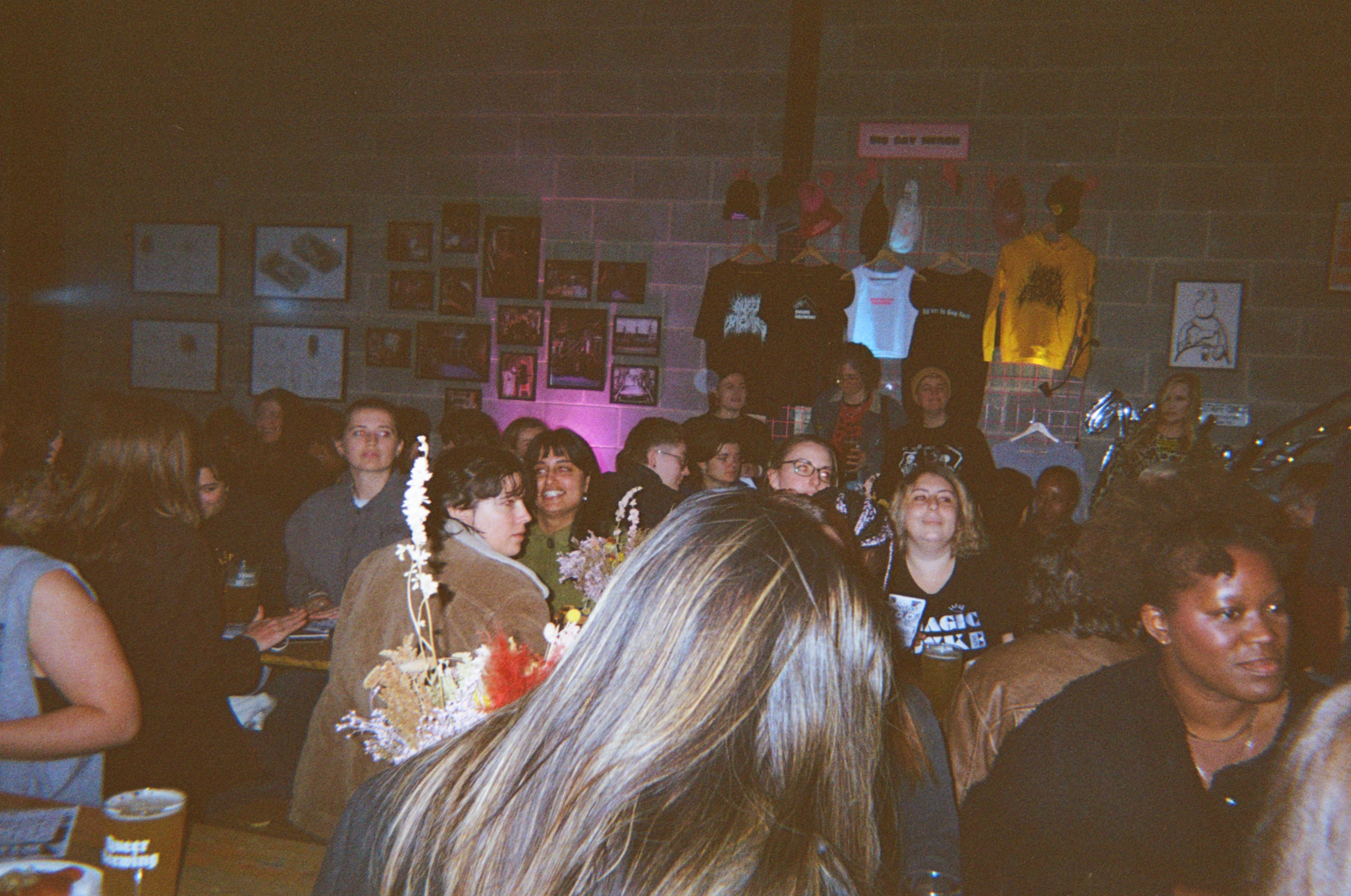 Crowded indoor venue with people gathered, warm lighting, posters on walls, blonde hair in foreground, dimly lit atmosphere.