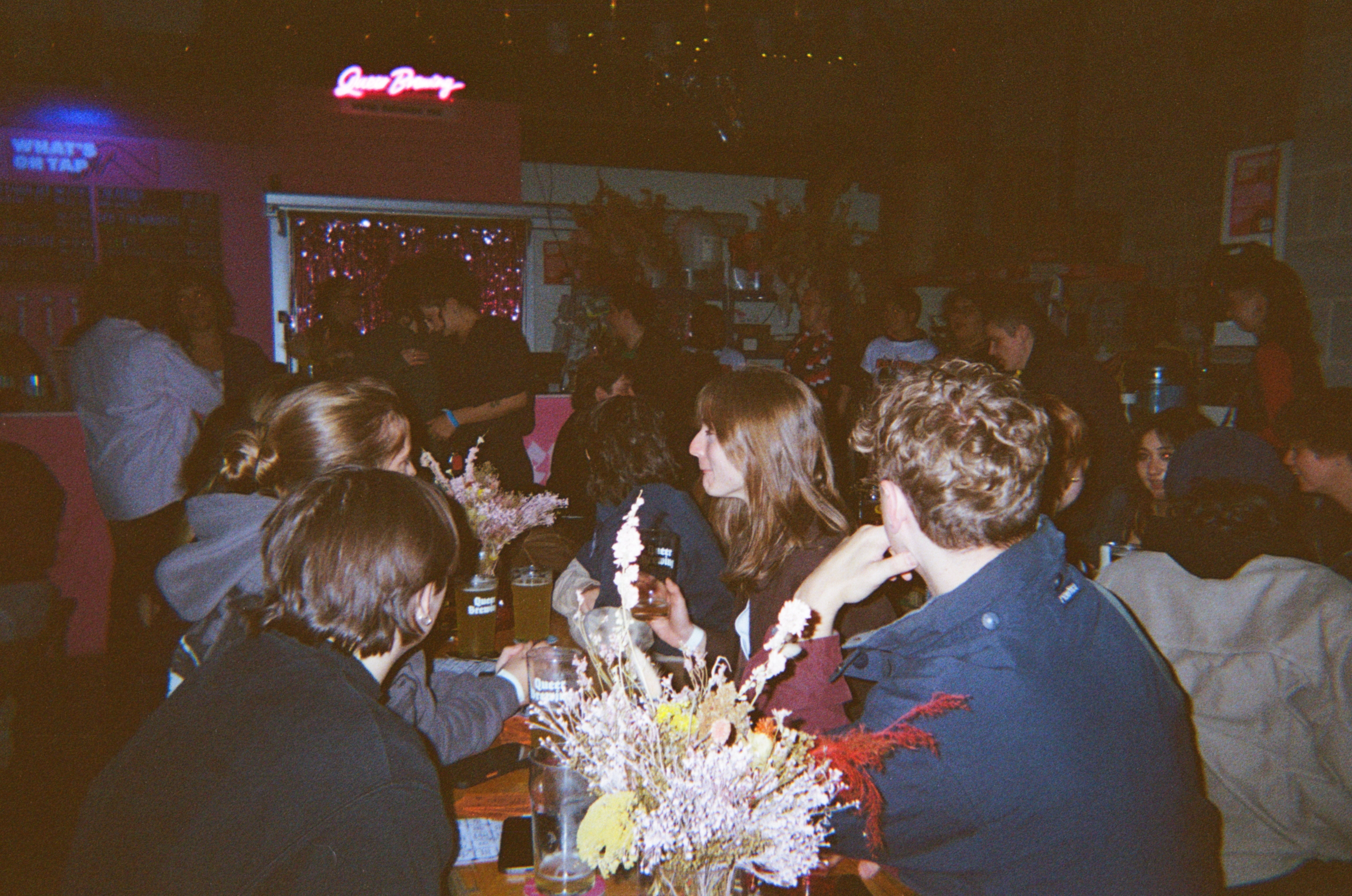 People gathered around table with flowers in dimly lit restaurant interior, red neon sign visible on wall in background.