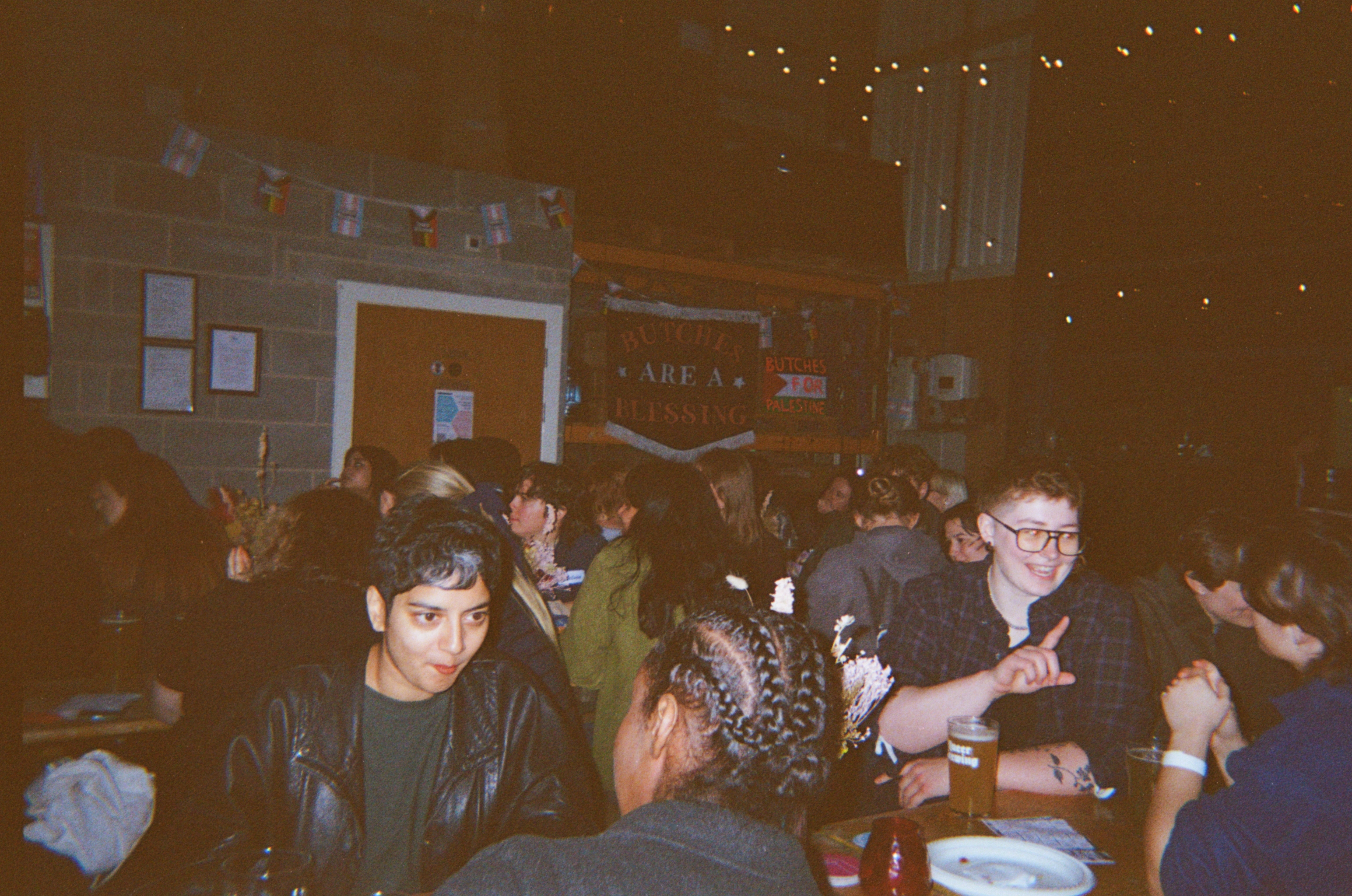 Group of young people sitting at tables in dimly lit pub or restaurant with warm brown lighting and blurred background.