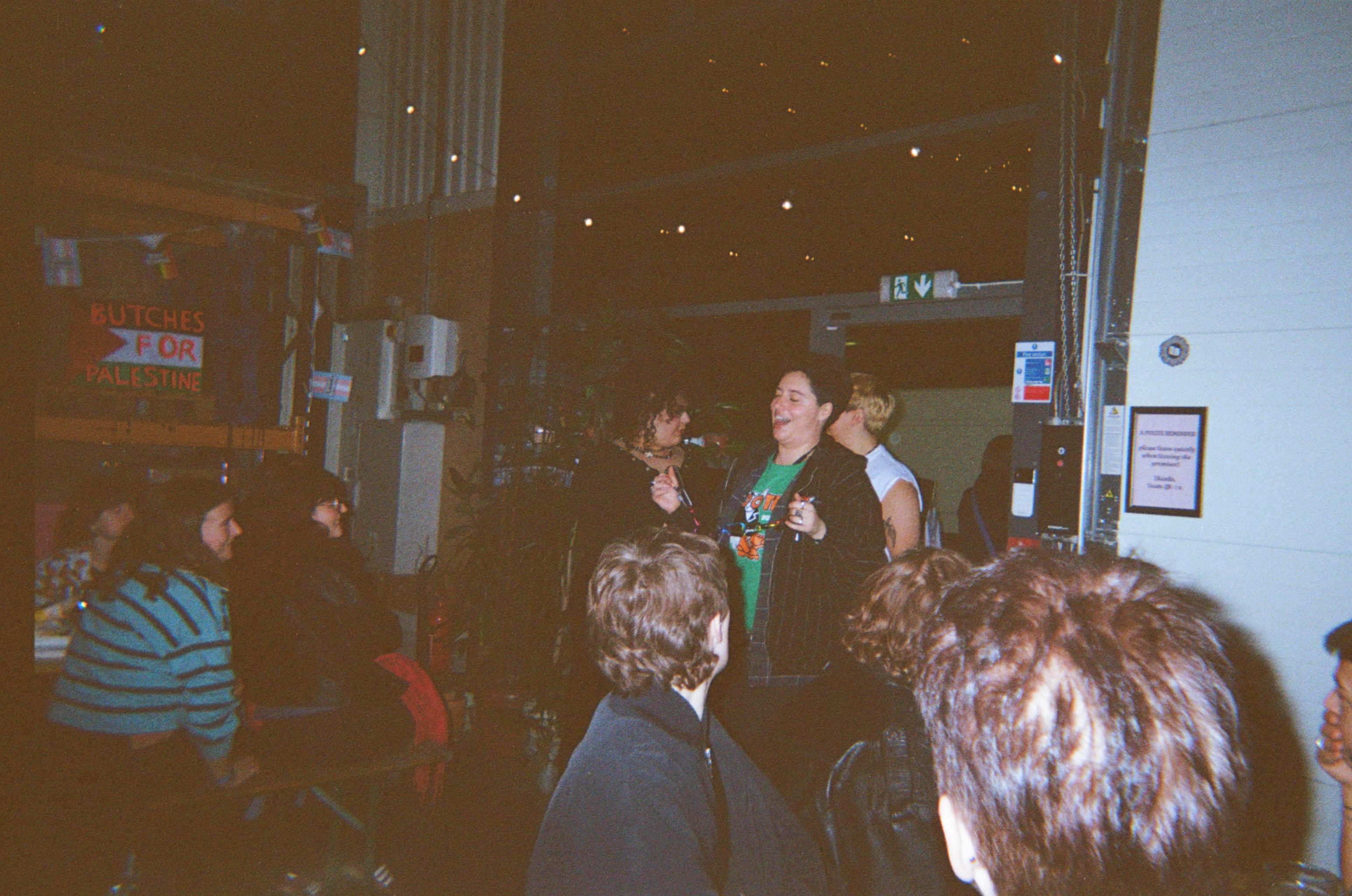 Two people standing behind a bar counter in dim lighting, with patrons' heads visible in foreground. Warm brown tones dominate the scene.