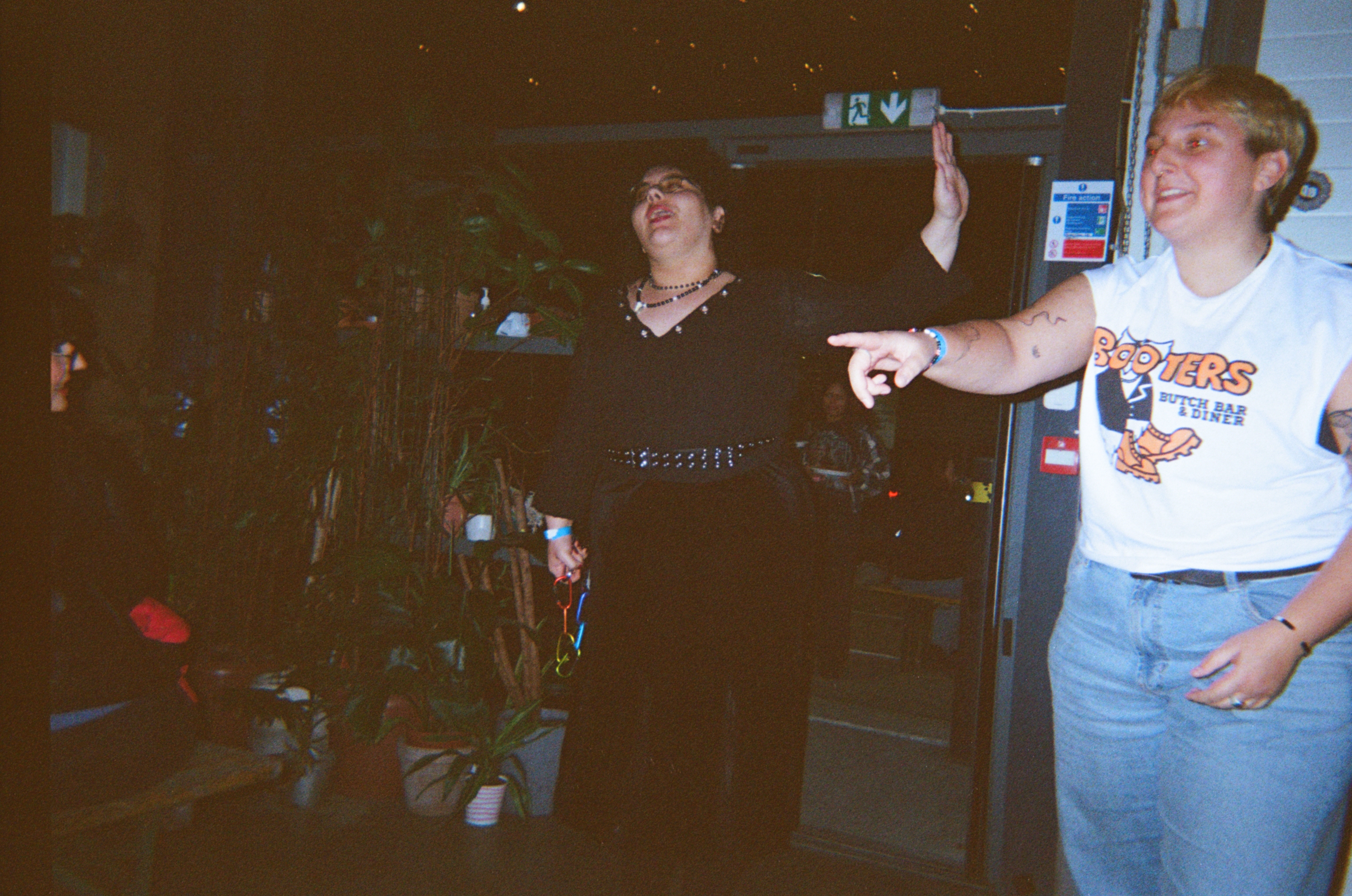 Two people playing arcade games in dimly lit venue. Person on right wears white t-shirt and jeans, person on left in dark clothing.