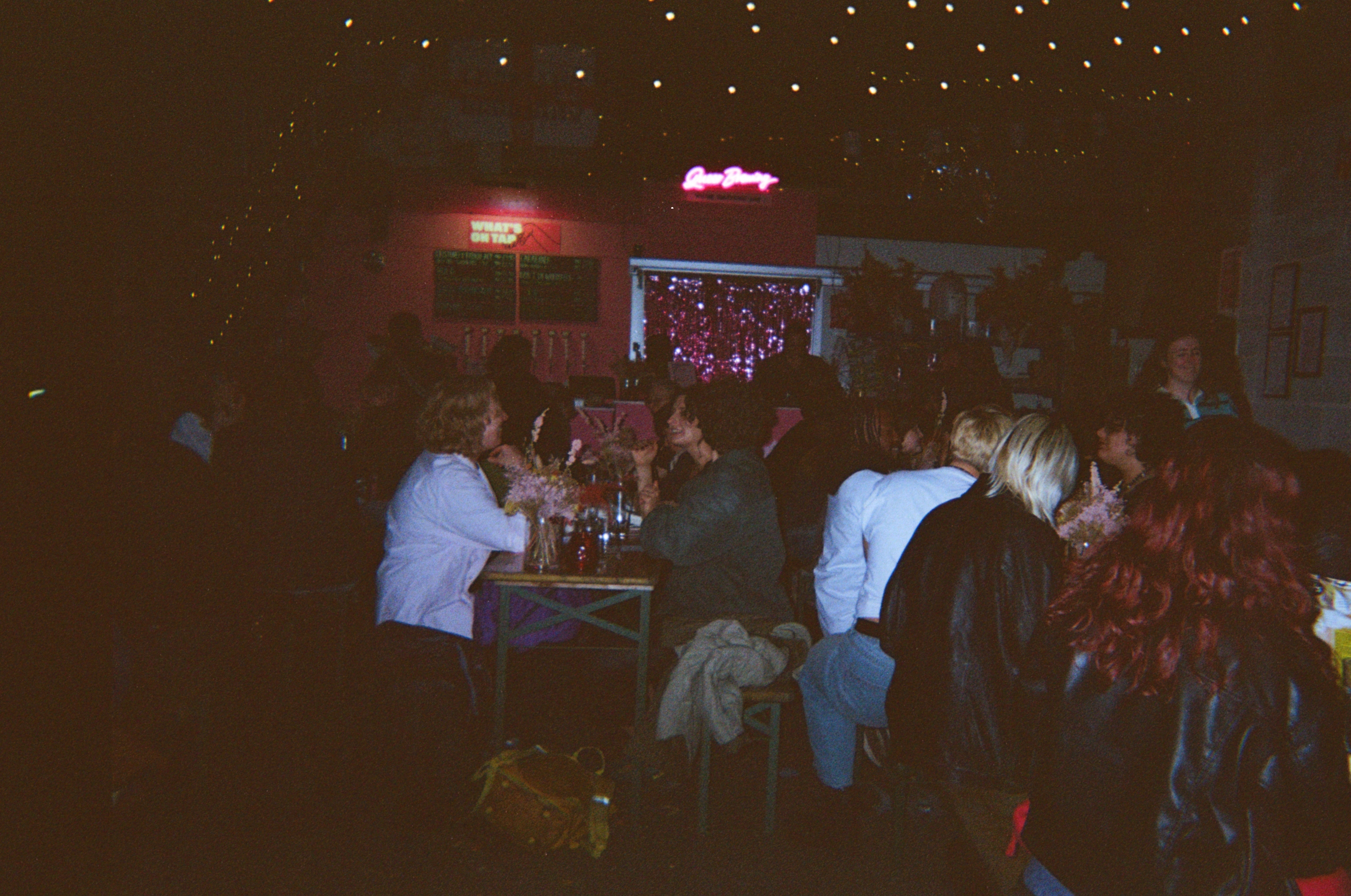 Dimly lit pub interior with people gathered around tables, red neon signs glowing on dark walls, warm orange lighting throughout.
