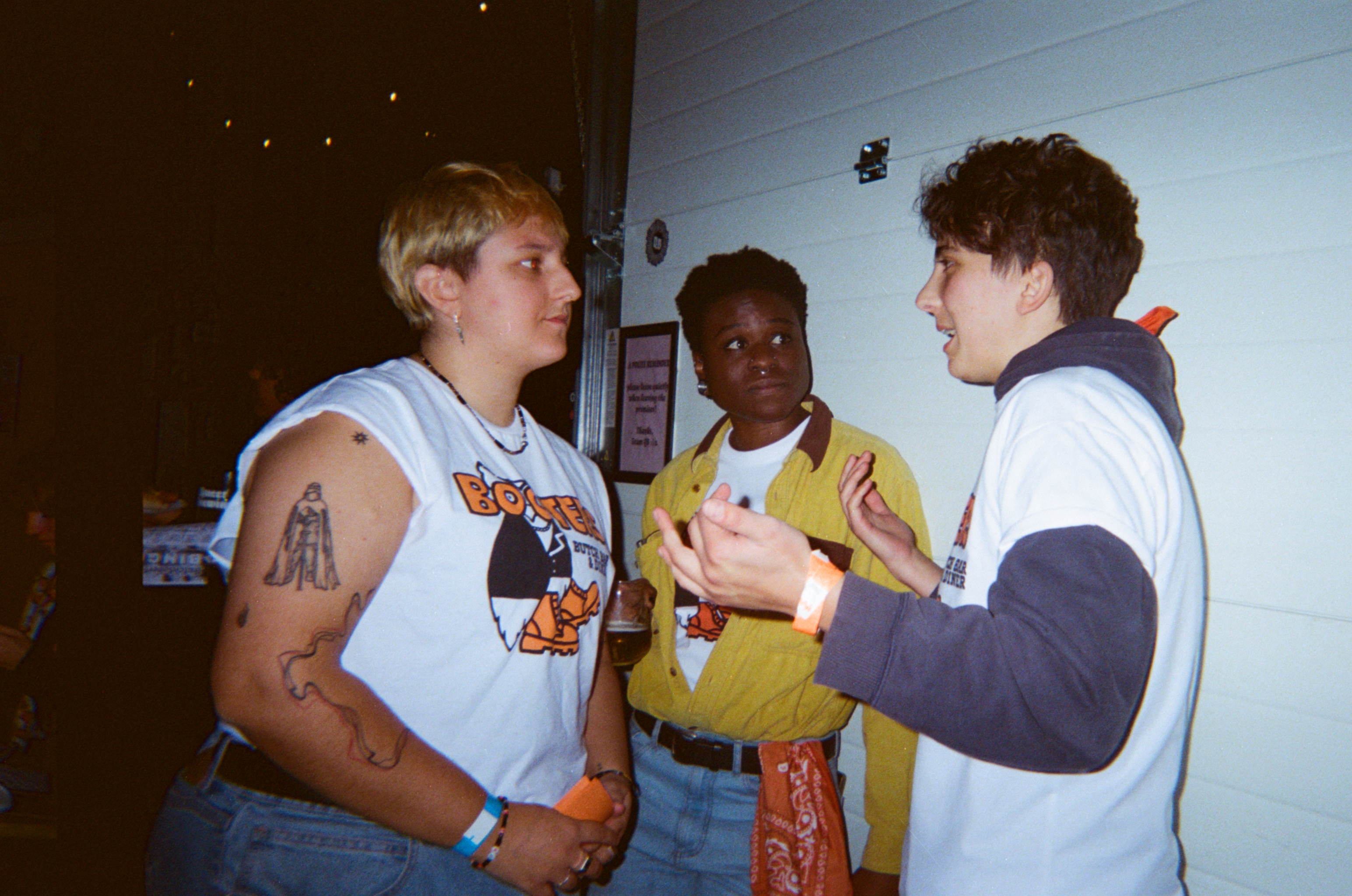 Three teenage boys chatting indoors. Left boy wears light blue sleeveless shirt, middle boy in yellow top, right boy in dark tracksuit top.